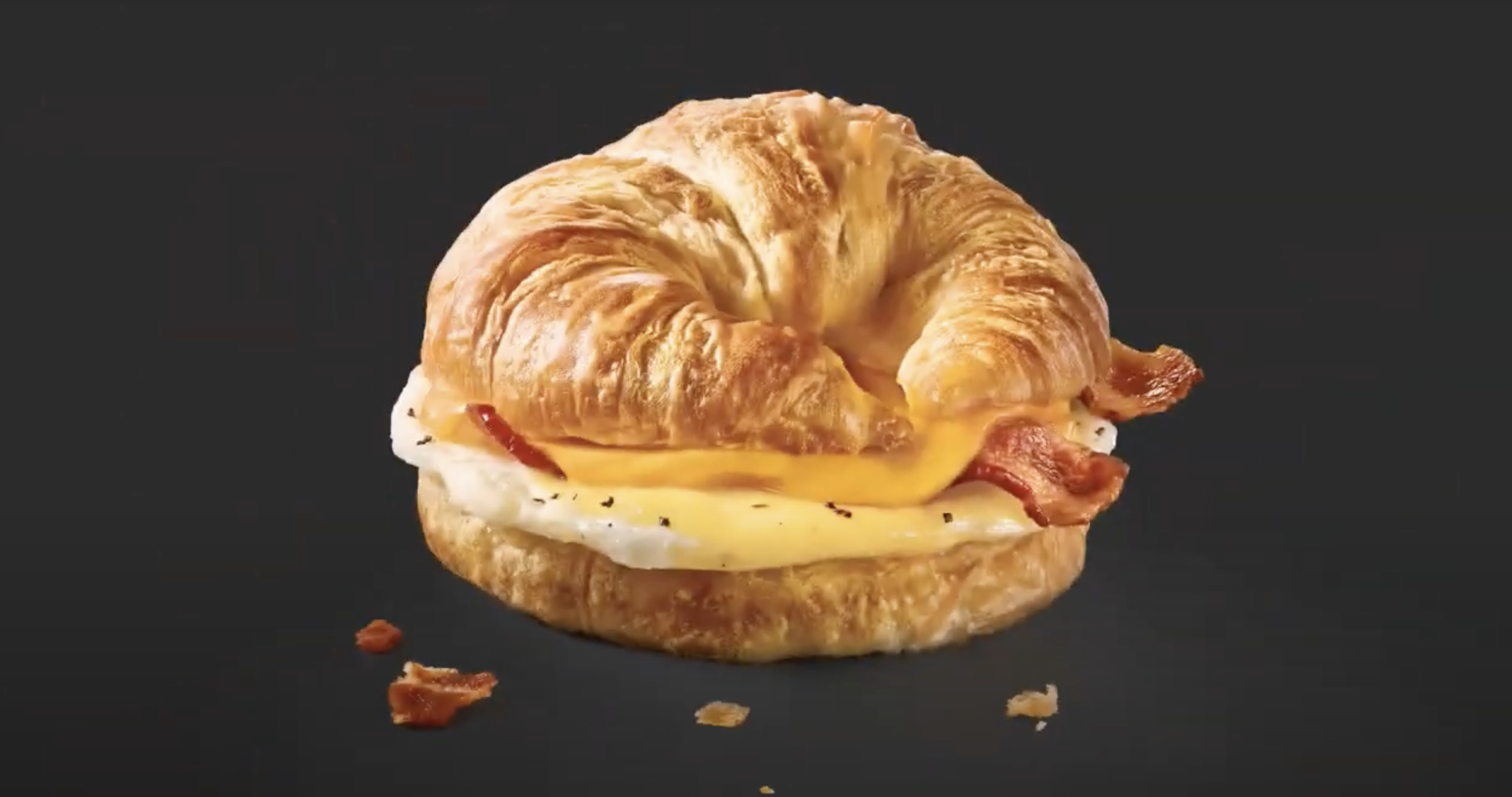Egg and bacon breakfast sandwich with a croissant, fried egg, melted cheese, and bacon on a black background.