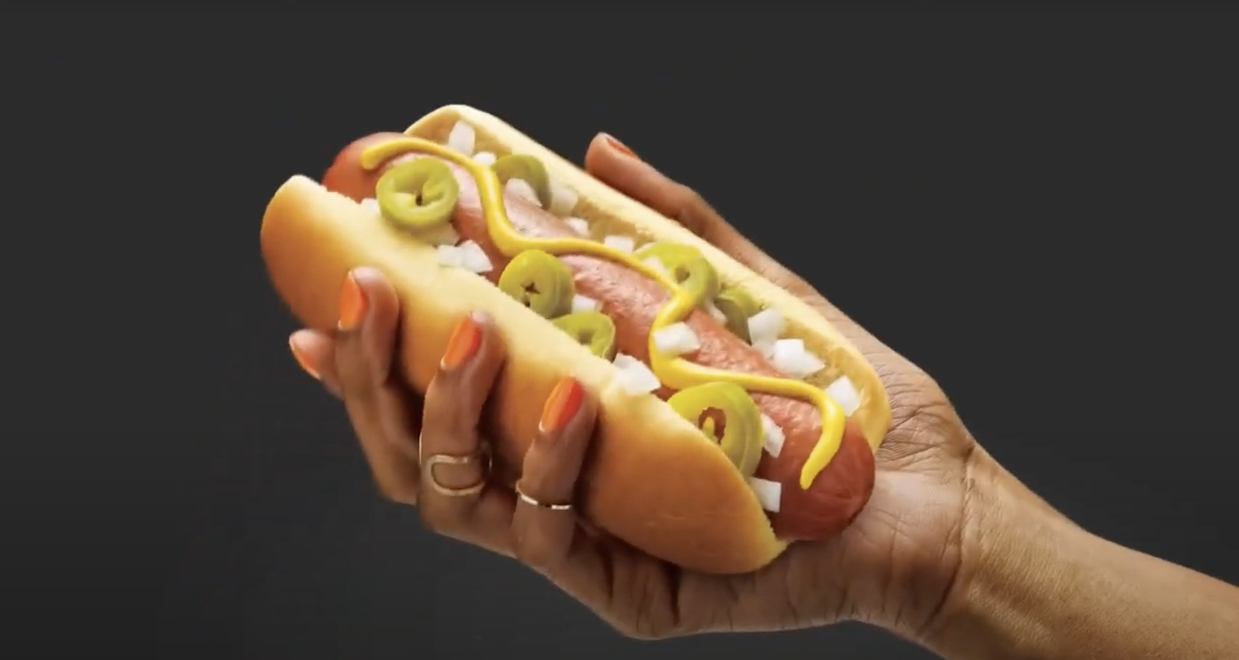 A person holding a hot dog in a bun, topped with sliced green jalapeños, diced onions, and yellow mustard against a dark background.