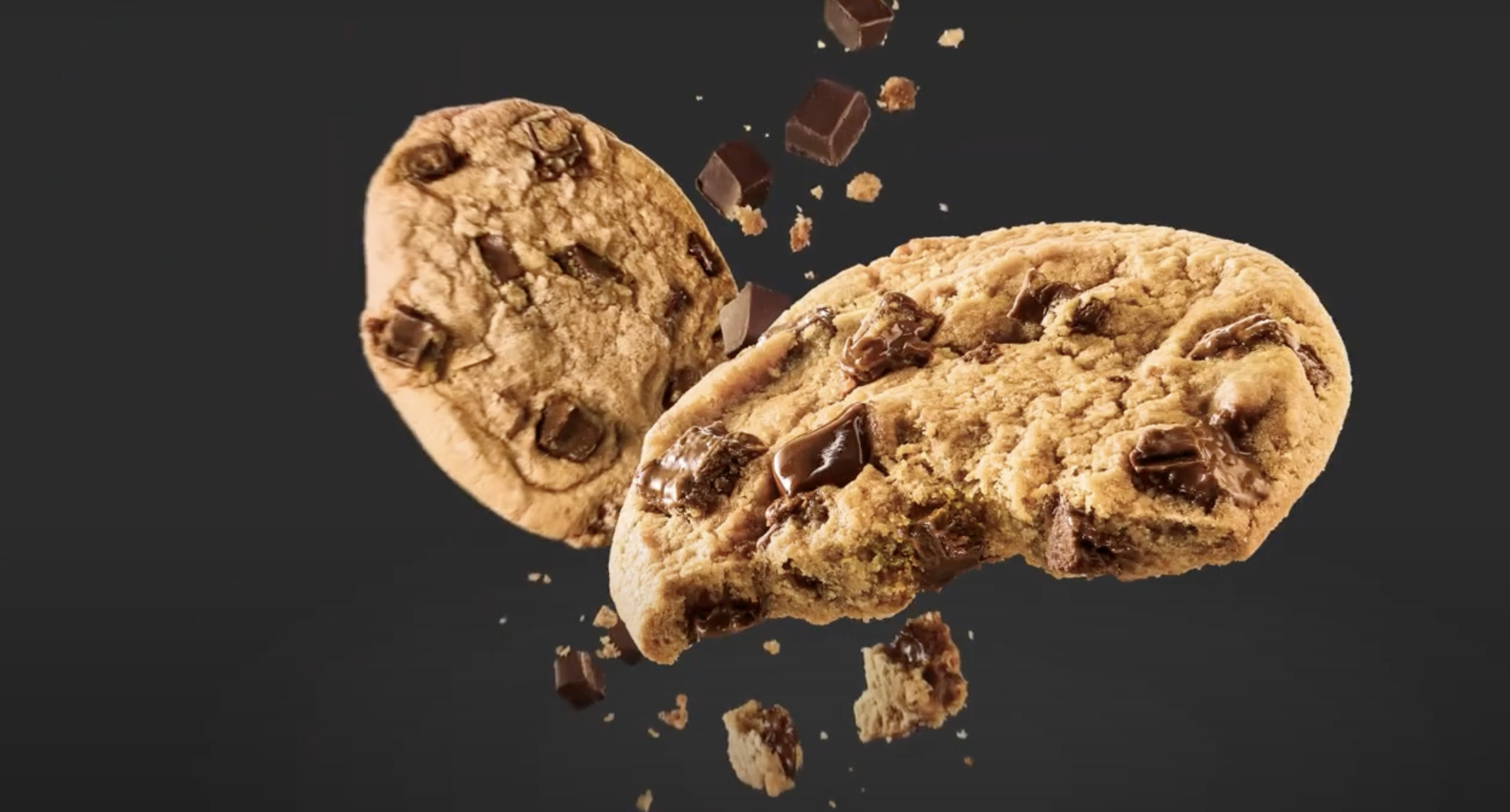 Two chocolate chip cookies breaking apart, with chocolate chunks and crumbs flying in the air against a dark background.