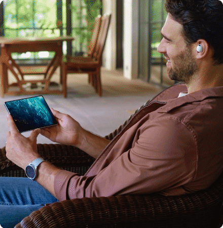 A man sitting on a wicker chair indoors, holding a smartphone with a scenic outdoor view on the screen. He is smiling, wearing wireless earbuds, a watch, and a brown shirt.