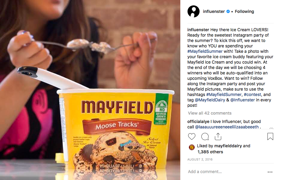 Close-up of a container of Mayfield Moose Tracks ice cream with a spoon inserted, with a person in the background holding a spoonful of ice cream.