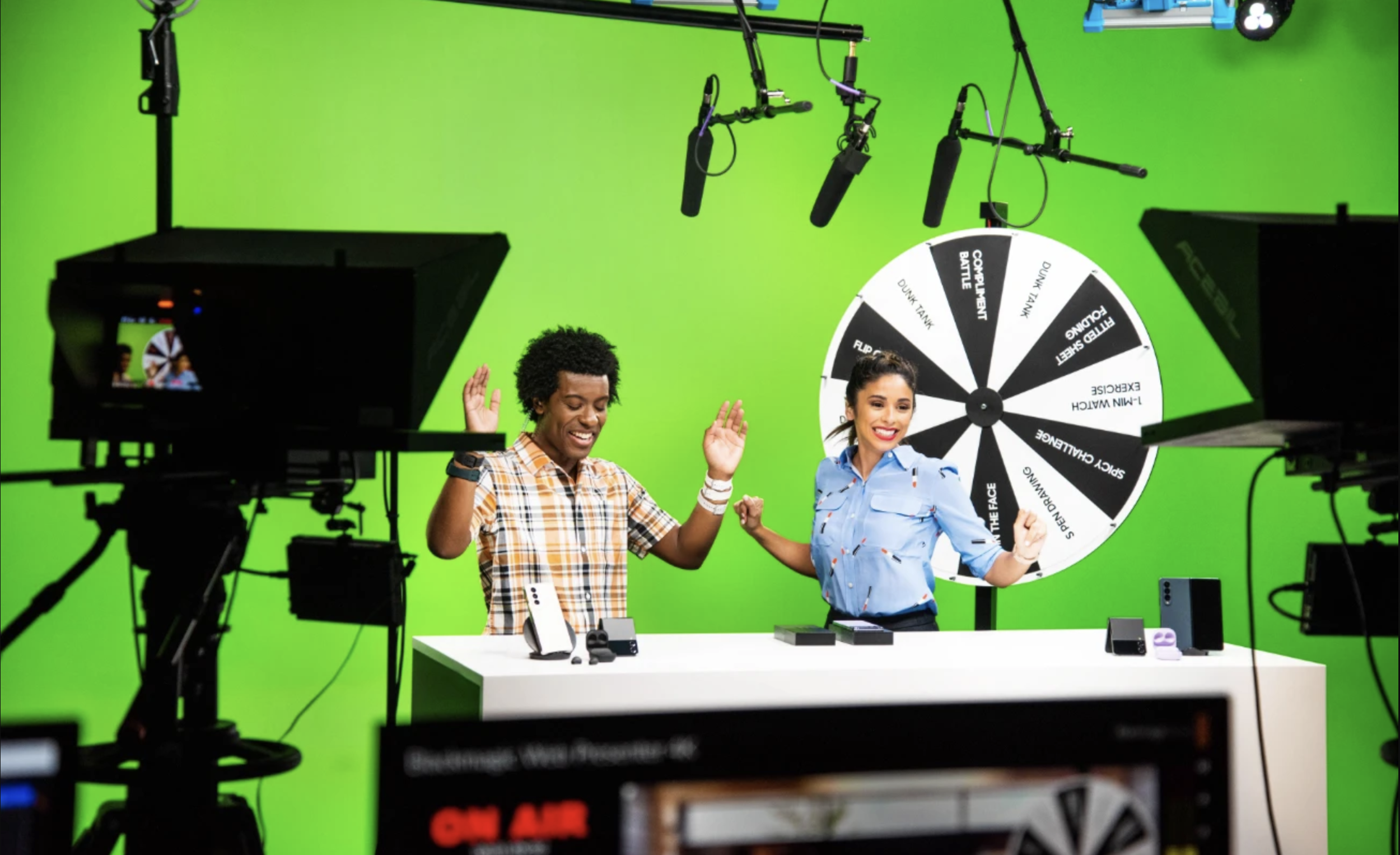 Two women hosting a game show in front of a green screen, with a spinning wheel and recording equipment around them.