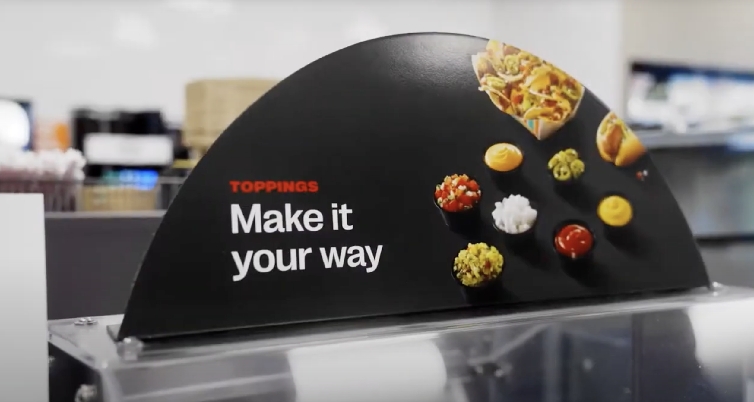 A black sign with red and white text that reads 'TOPPINGS Make it your way' and displays small bowls of various food toppings such as chopped vegetables, sauces, and condiments.