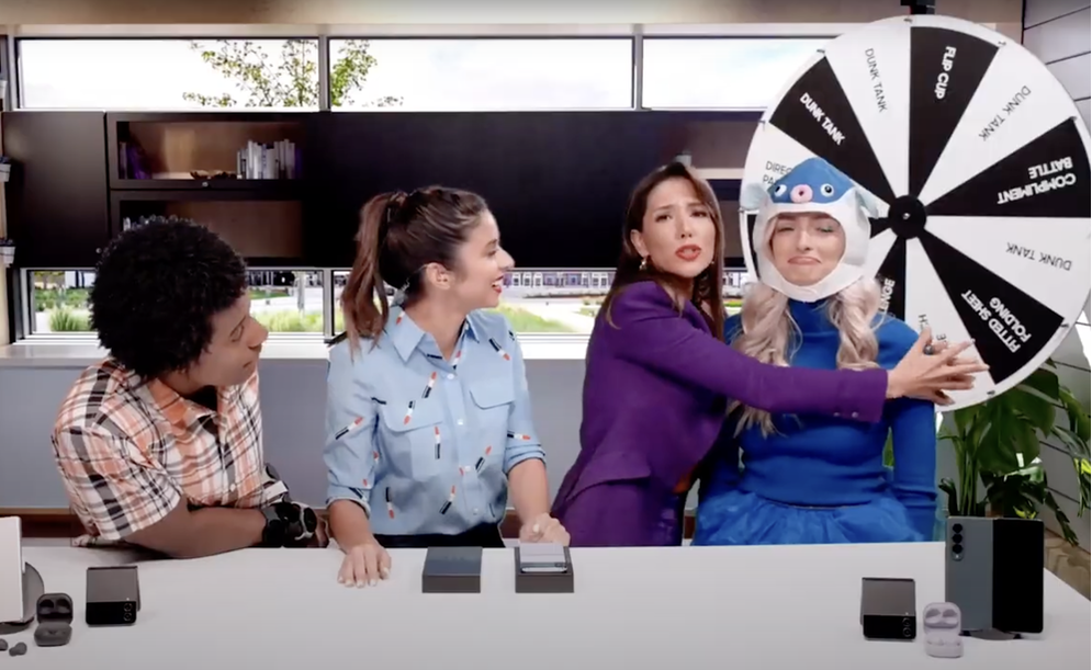 Four women sitting at a table, one wearing a fun shark mask on a spinning wheel, with the wheel labeled with sections like 'Re-Spin,' 'DUNK TANK,' and 'COUPON LIFT,' in a modern office space.