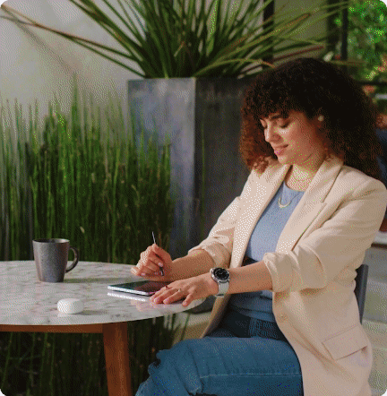A woman with curly hair sitting at a marble table in a bright indoor space, writing in a notebook with a pen, with a mug and a small container on the table, and large green plants in the background.