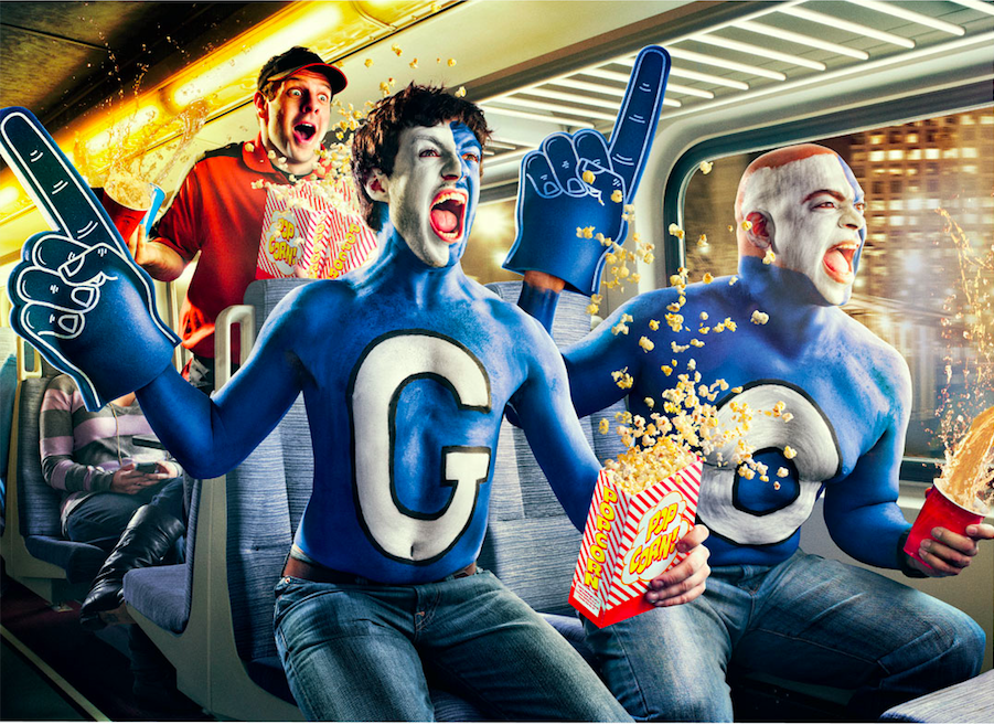 Three people dressed as comics fans celebrating and cheering while sitting on a train. The two in the front are painted blue with big letters 'G' and 'O' on their bodies, holding popcorn and soda. They are wearing large foam gloves and costumes, with one having face paint resembling a comic character. The person in the back is wearing a red shirt and holding popcorn, looking excited.