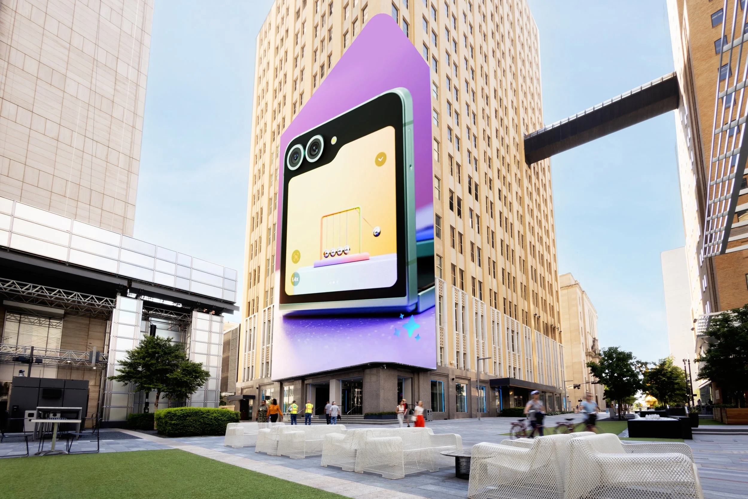 Large digital billboard on a city building displaying a colorful advertisement for a smartphone with a dual camera and a playful gaming scene. Pedestrians walking and cyclists riding through a modern urban plaza with benches, trees, and surrounding high-rise buildings under a clear blue sky.