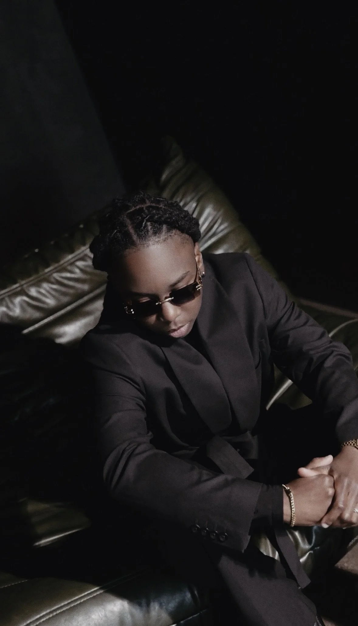 A man dressed in a black suit and sunglasses is sitting in a black leather chair with a metallic back in a dimly lit room.