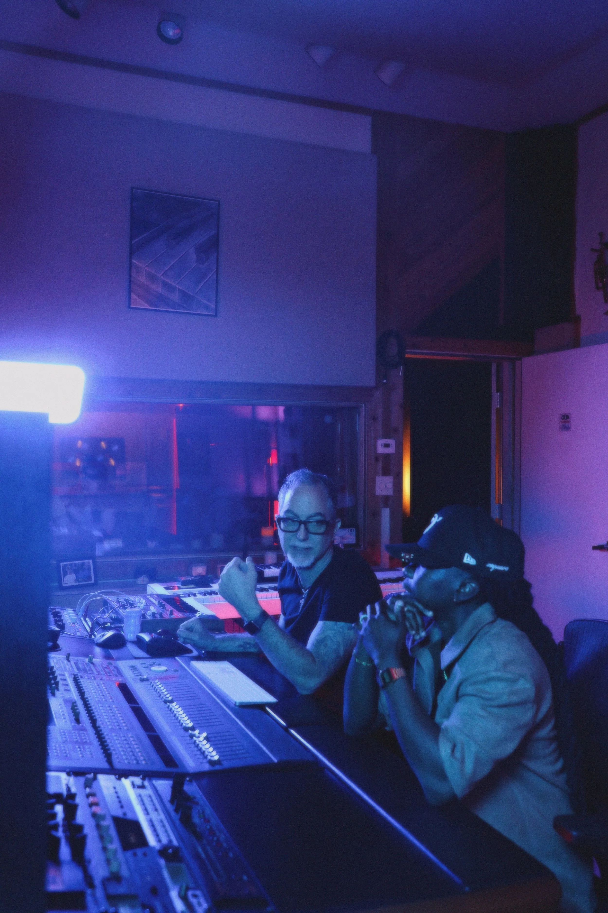 Two men are sitting in a recording studio with audio equipment and a mixing console, illuminated by blue and purple lighting.