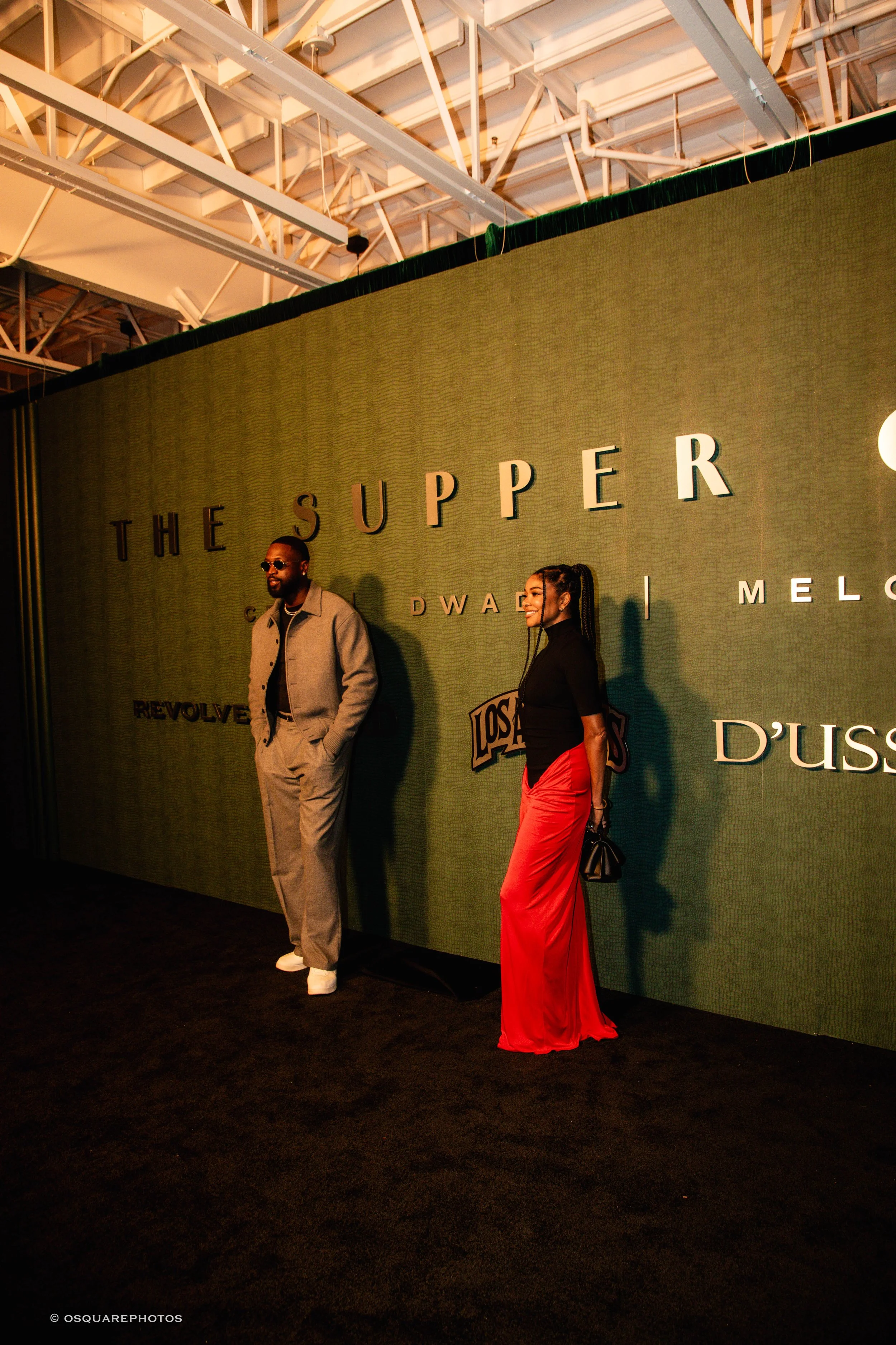 Dwyane Wade and Gabrielle Union posing on the black carpet in front of “The Supper Club” backdrop during NBA All-Star Weekend, under warm event lighting.