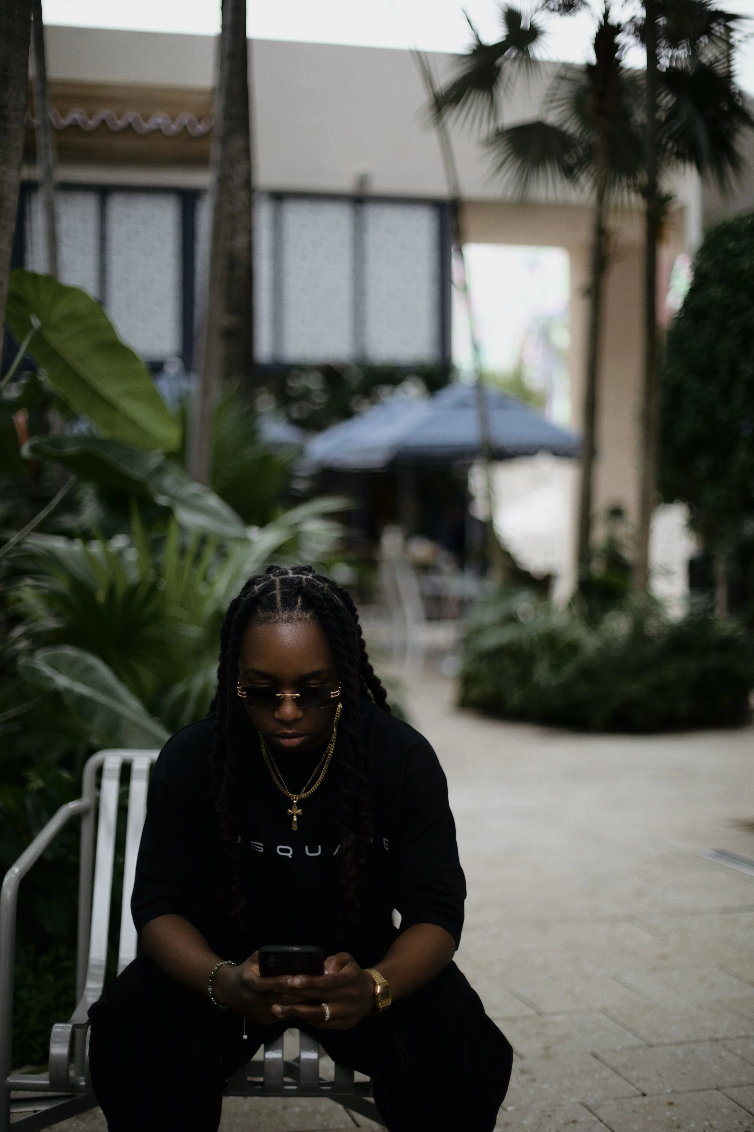 Person wearing black OSQUARE shirt sitting on a bench in a tropical courtyard, looking down at their phone, surrounded by palm trees and greenery.