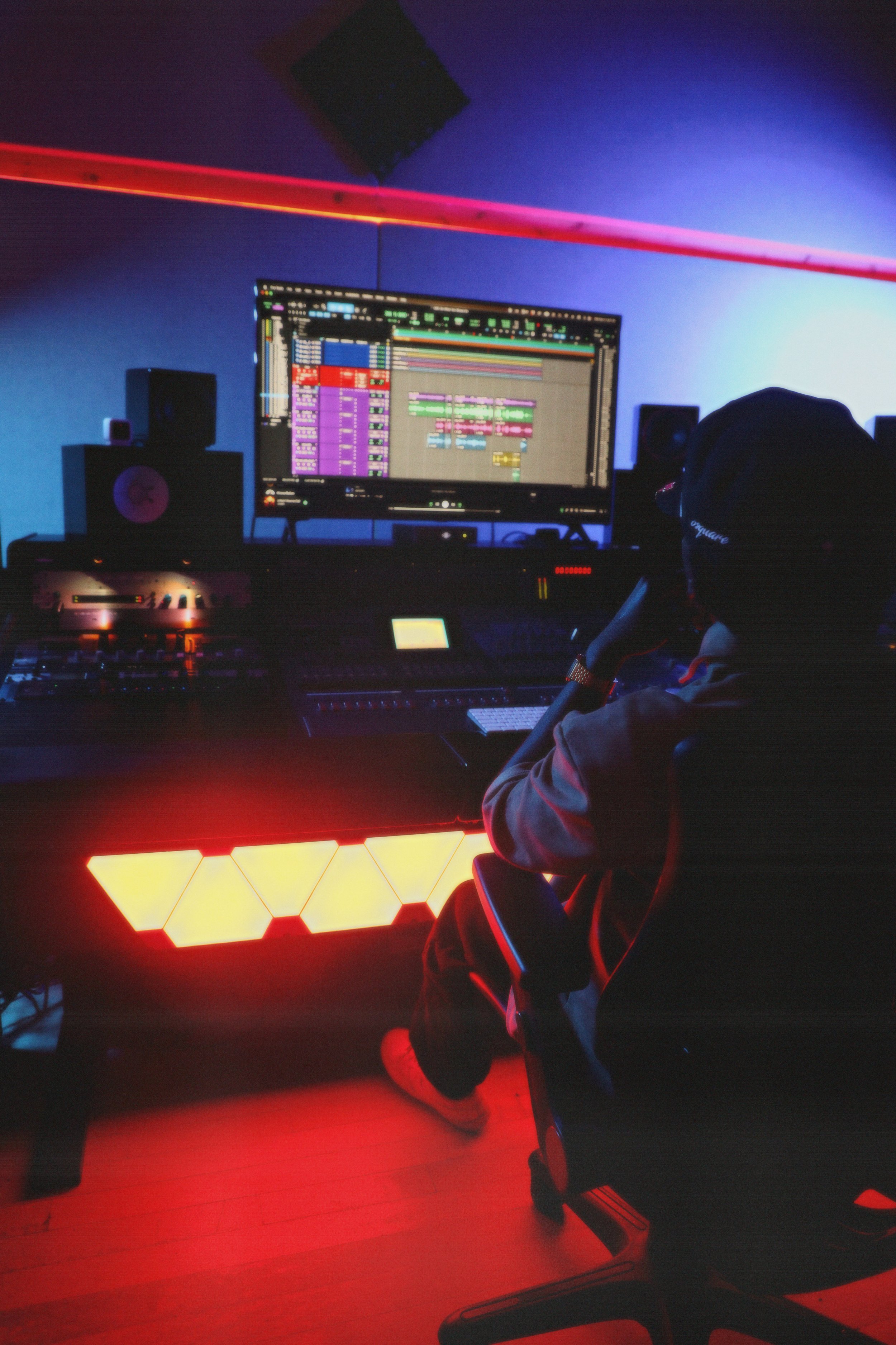 A music production studio with a large computer monitor displaying a colorful digital audio workstation. Black studio speakers are positioned on either side of the monitor. A person is seated at the desk wearing a hat and headphones, working on music. The room has vibrant lighting, with a red hexagonal light display on the desk and a neon red strip along the ceiling.