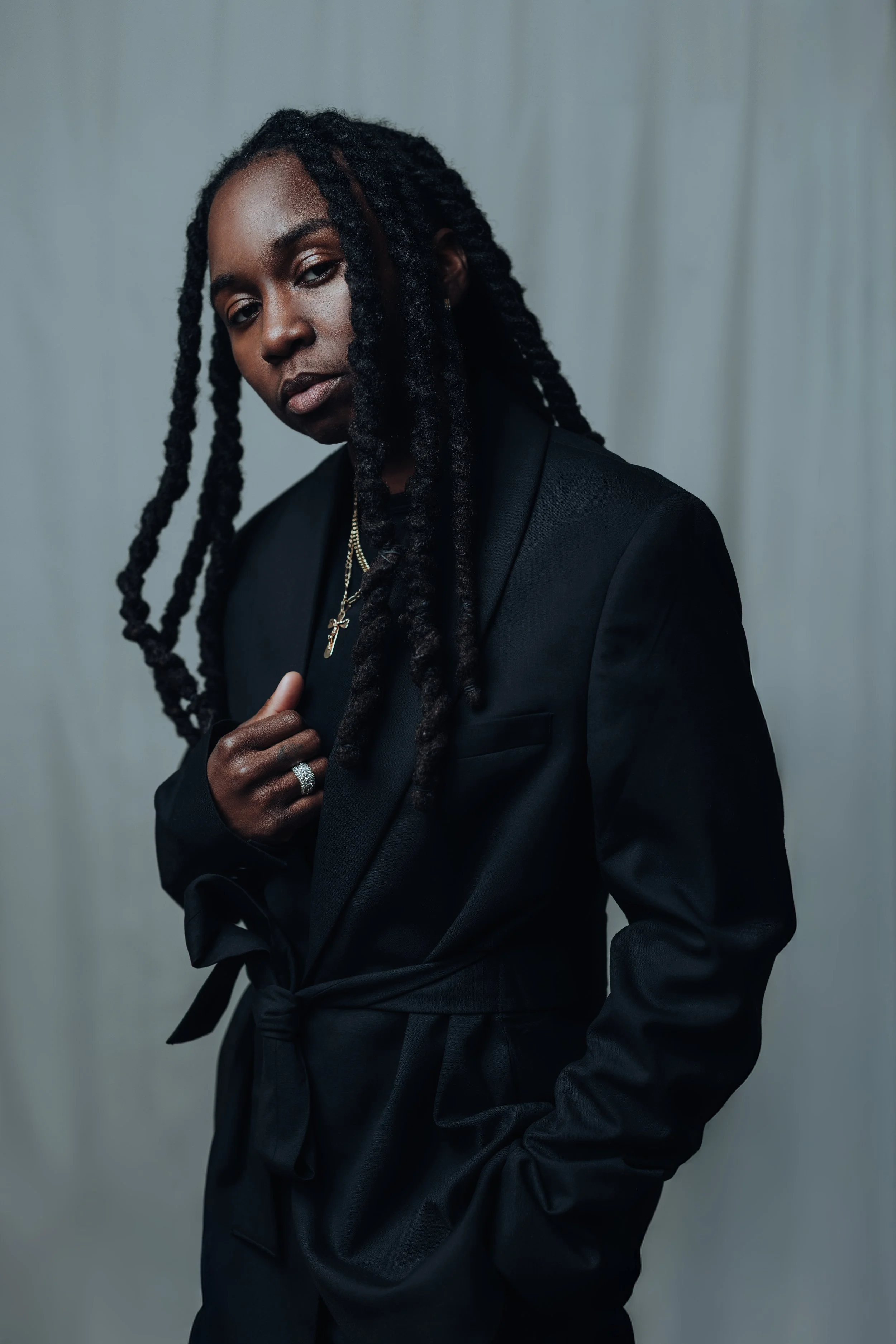 Portrait of a Black artist wearing a tailored black suit with a tied waist belt, layered gold necklaces, and long twisted locs, standing against a soft gray backdrop with a calm, confident expression.
