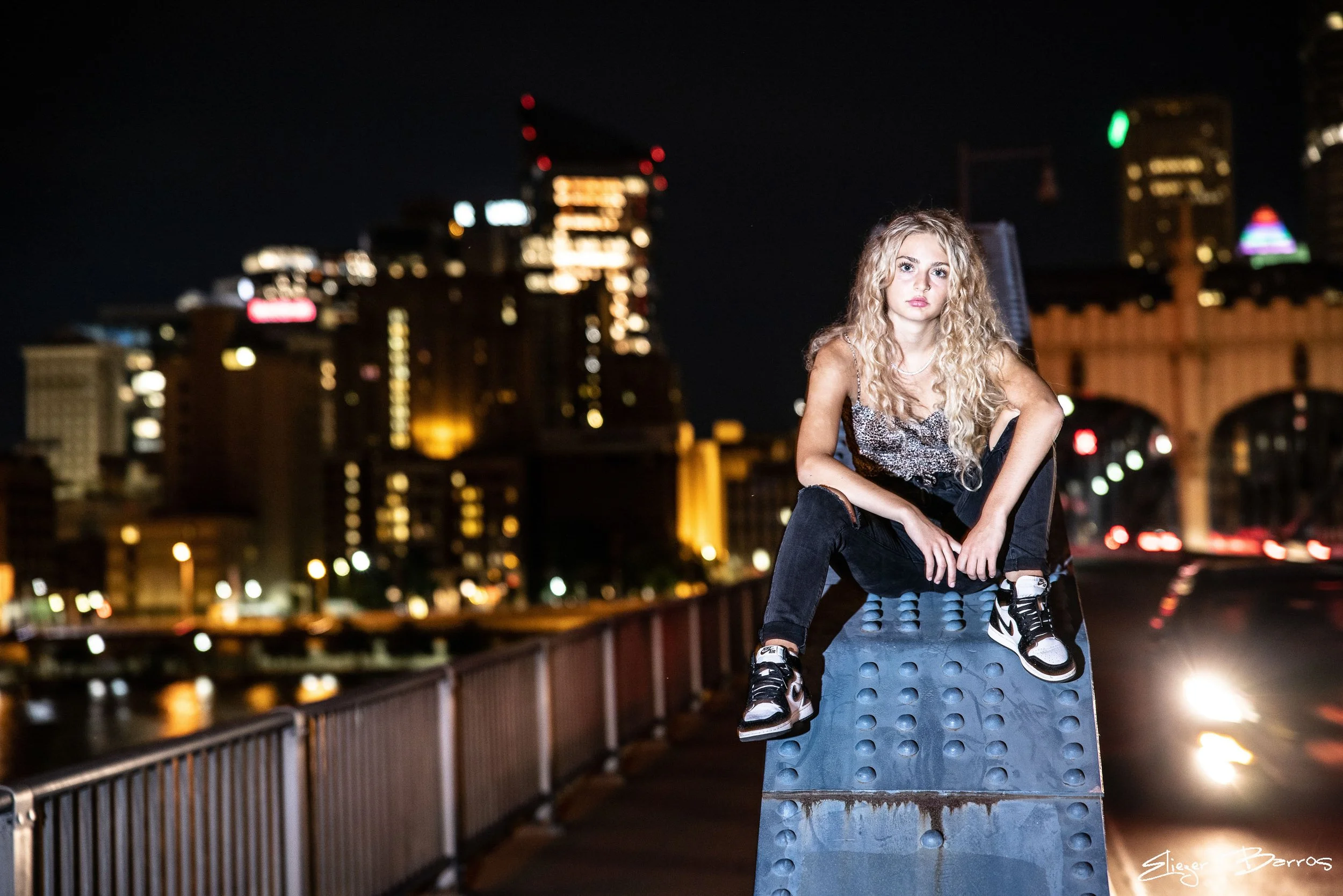 Senior Pictures Pittsburgh at Station Square and Smithfield Street Bridge. Young woman with curly blonde hair posing at night on the waterfront with glowing Pittsburgh city lights for vibrant urban senior pictures.
