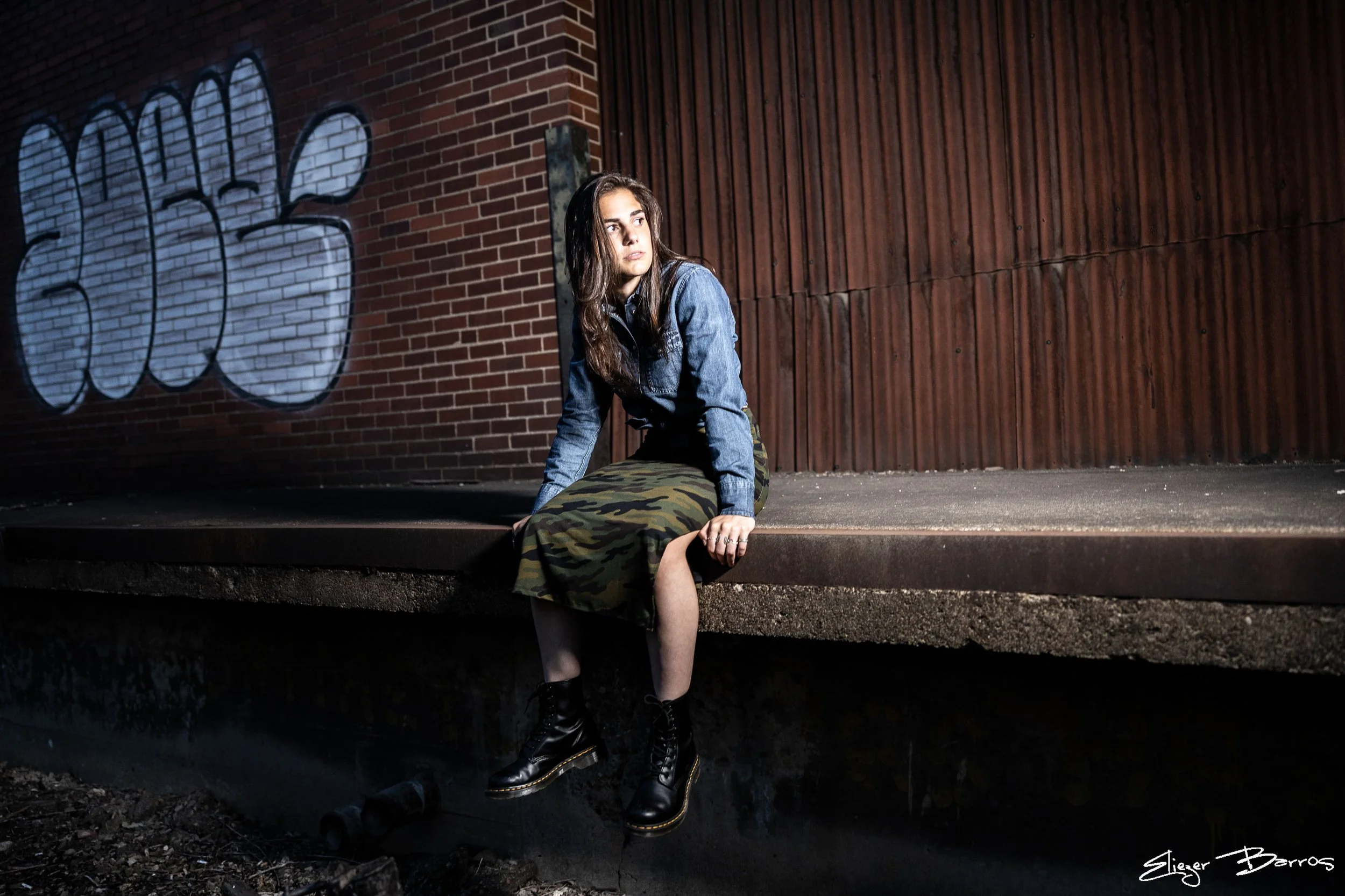 A young woman with long brown hair sitting on a ledge in front of a brick wall with graffiti, wearing a denim jacket, camo pants, and black combat boots.