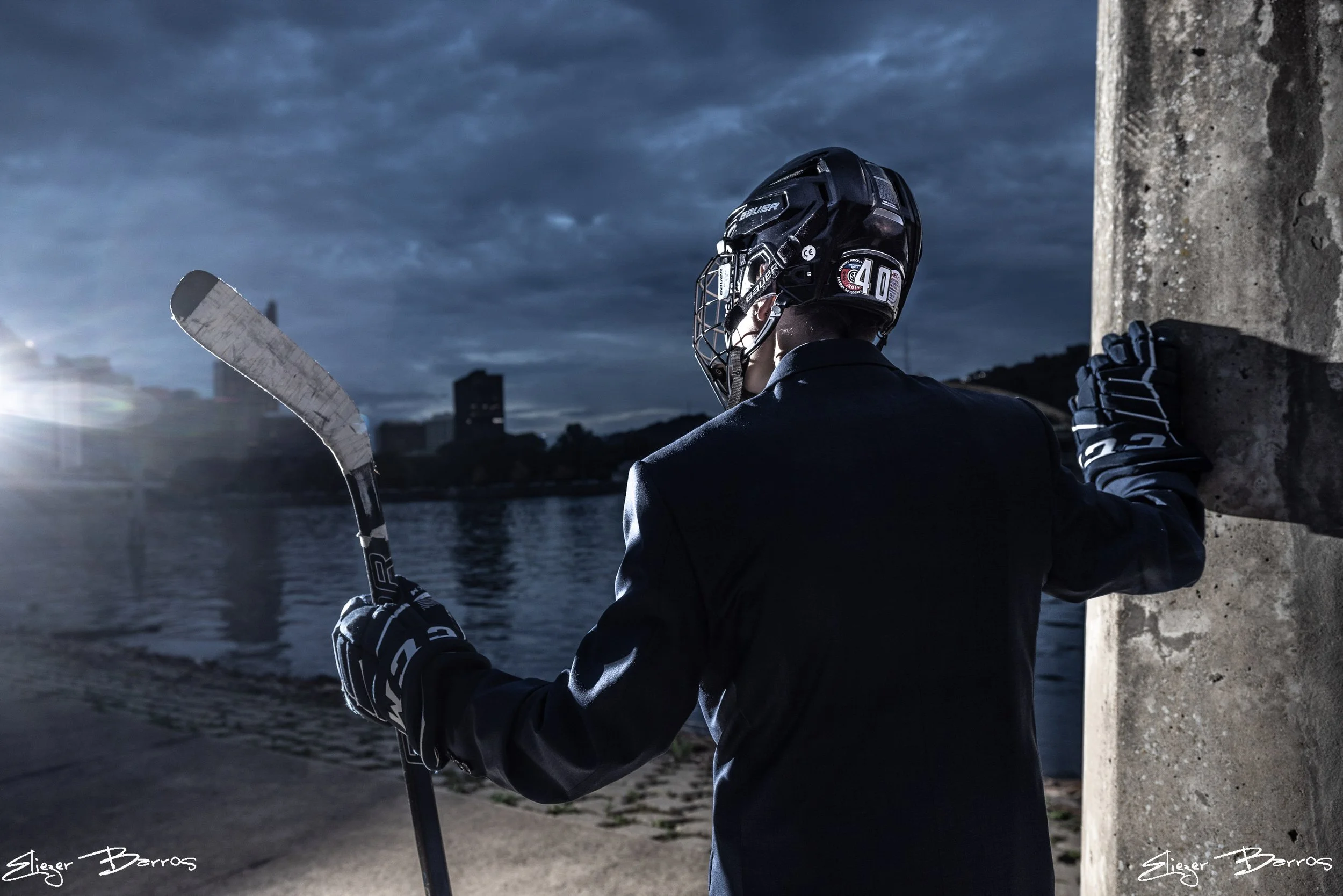 A person in hockey gear, including a helmet and gloves, is sitting on a concrete ledge near water under a cloudy sky, holding a hockey stick.