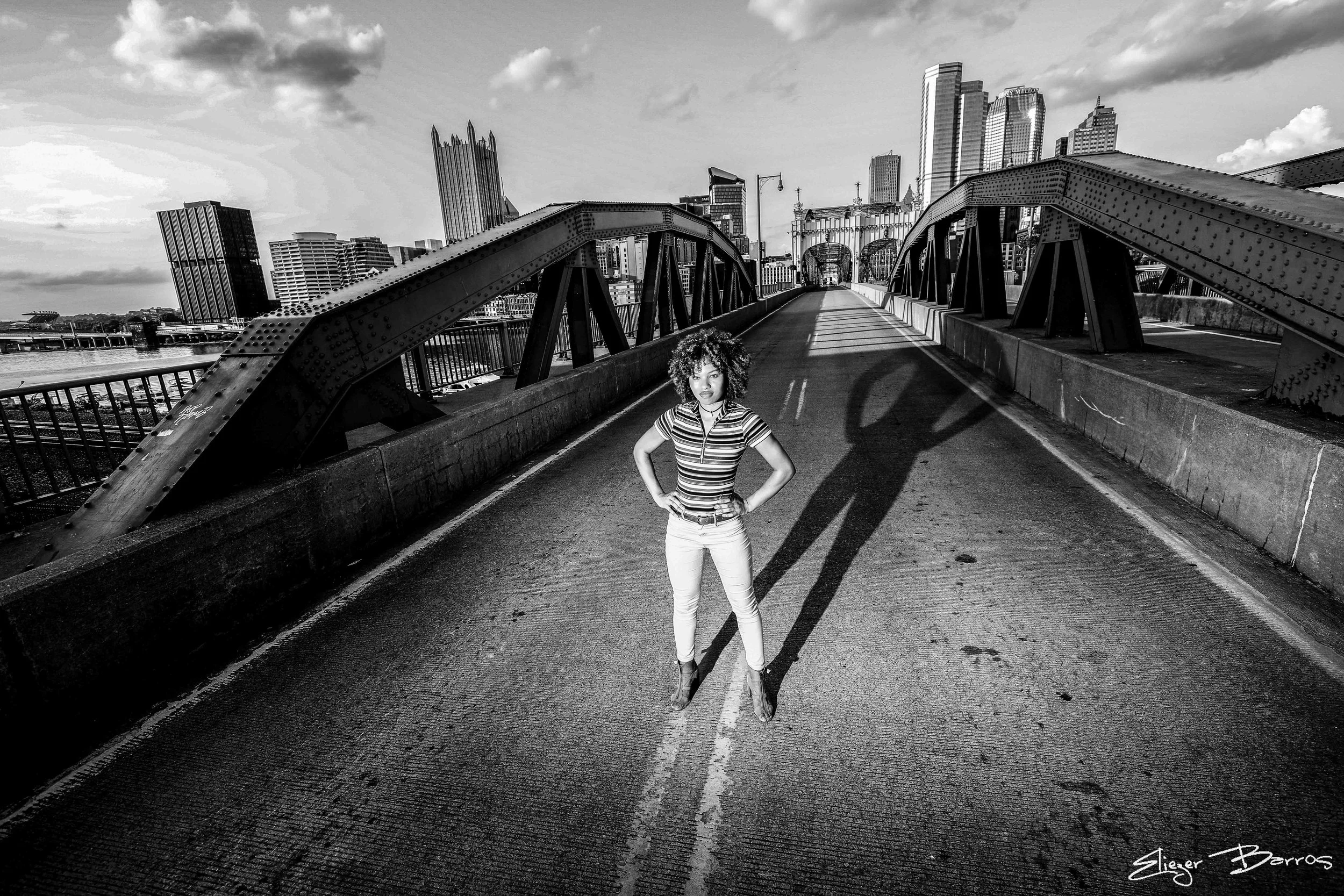 Black and white photo of a woman standing on a bridge in a cityscape, with tall skyscrapers in the background and shadows cast on the road.