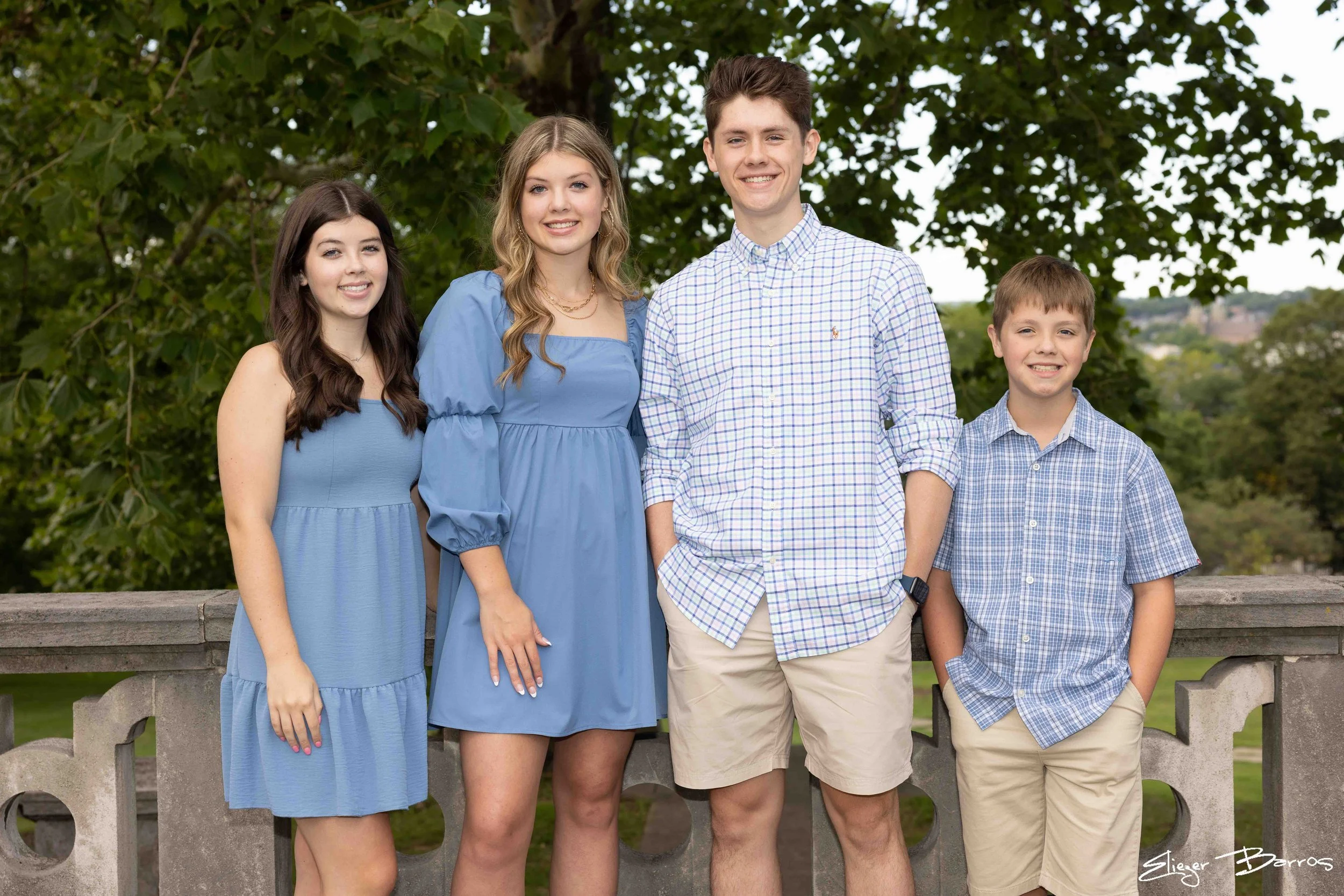 Four teenagers, two girls and two boys, standing outdoors in front of a leafy green tree, smiling at the camera. The girls are in blue dresses, and the boys are in light-colored shirts and khaki shorts.