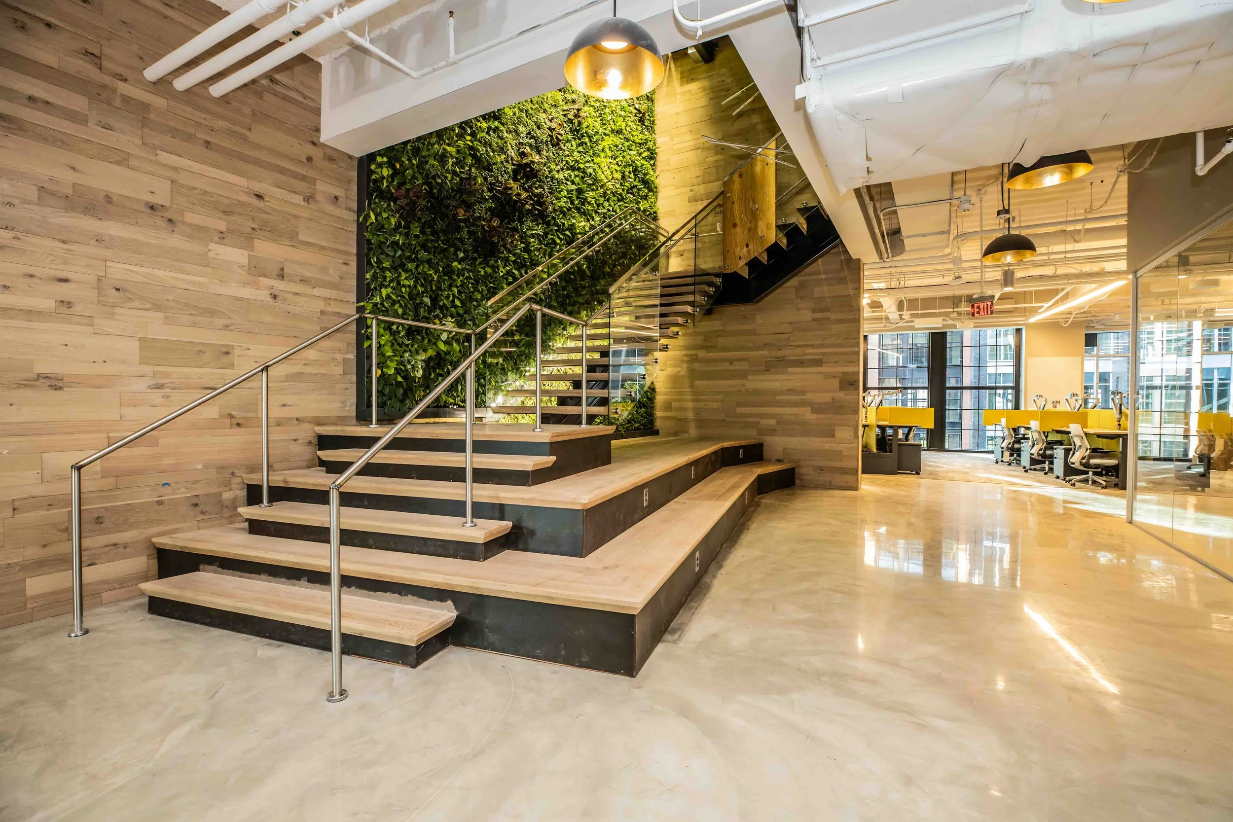 Interior view of a modern office space with wooden staircase, green wall, yellow desks, and large windows.