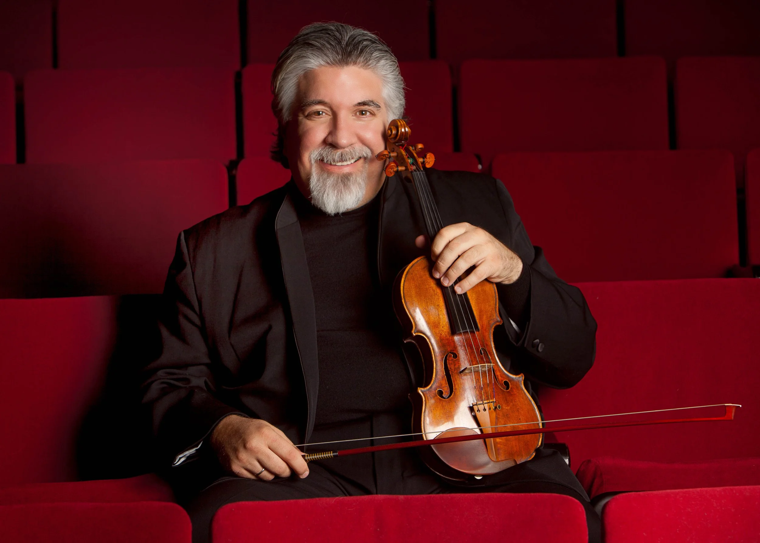 An older man with gray hair and a beard smiling while holding a violin and bow, sitting in an auditorium with red velvet seats.