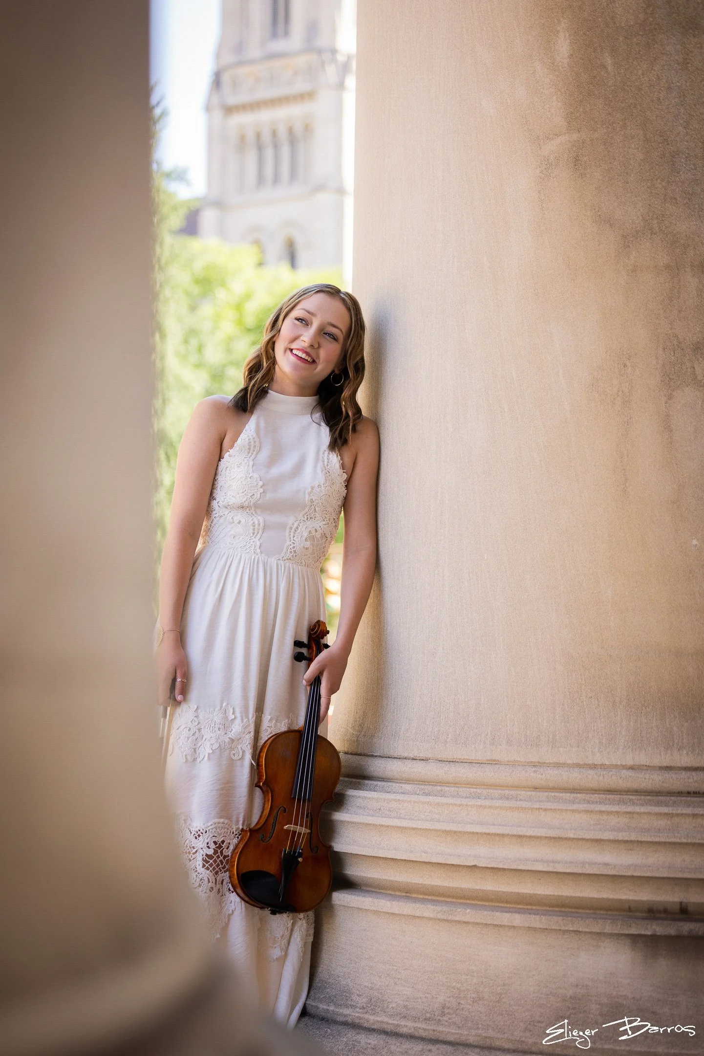 A young woman with shoulder-length hair, smiling, leaning against a beige wall outdoors during daytime, holding a violin in her right hand. She is wearing a sleeveless white lace dress. In the background, there are green trees and a building with cla