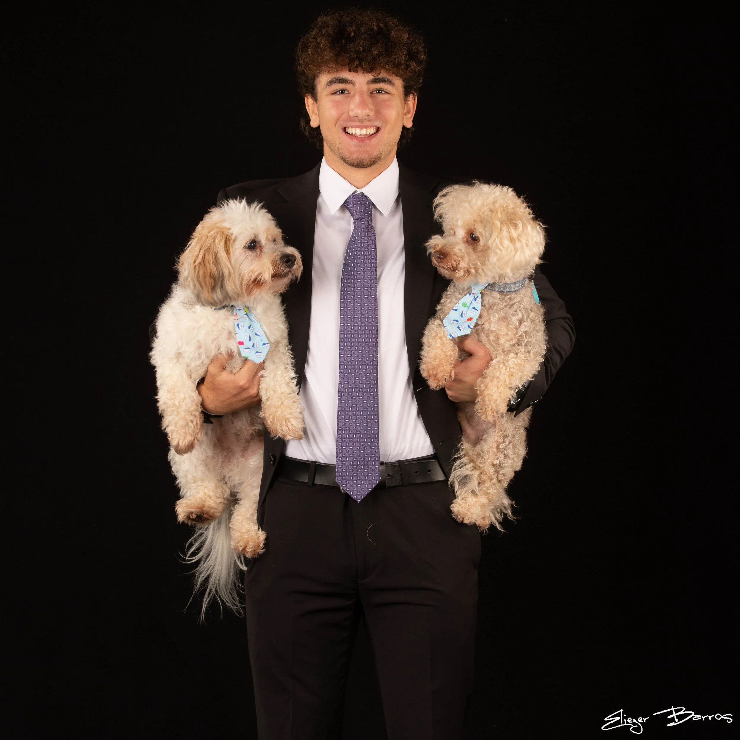 A young man in a suit and tie holding two small, fluffy, beige dogs with blue bandanas against a black background.