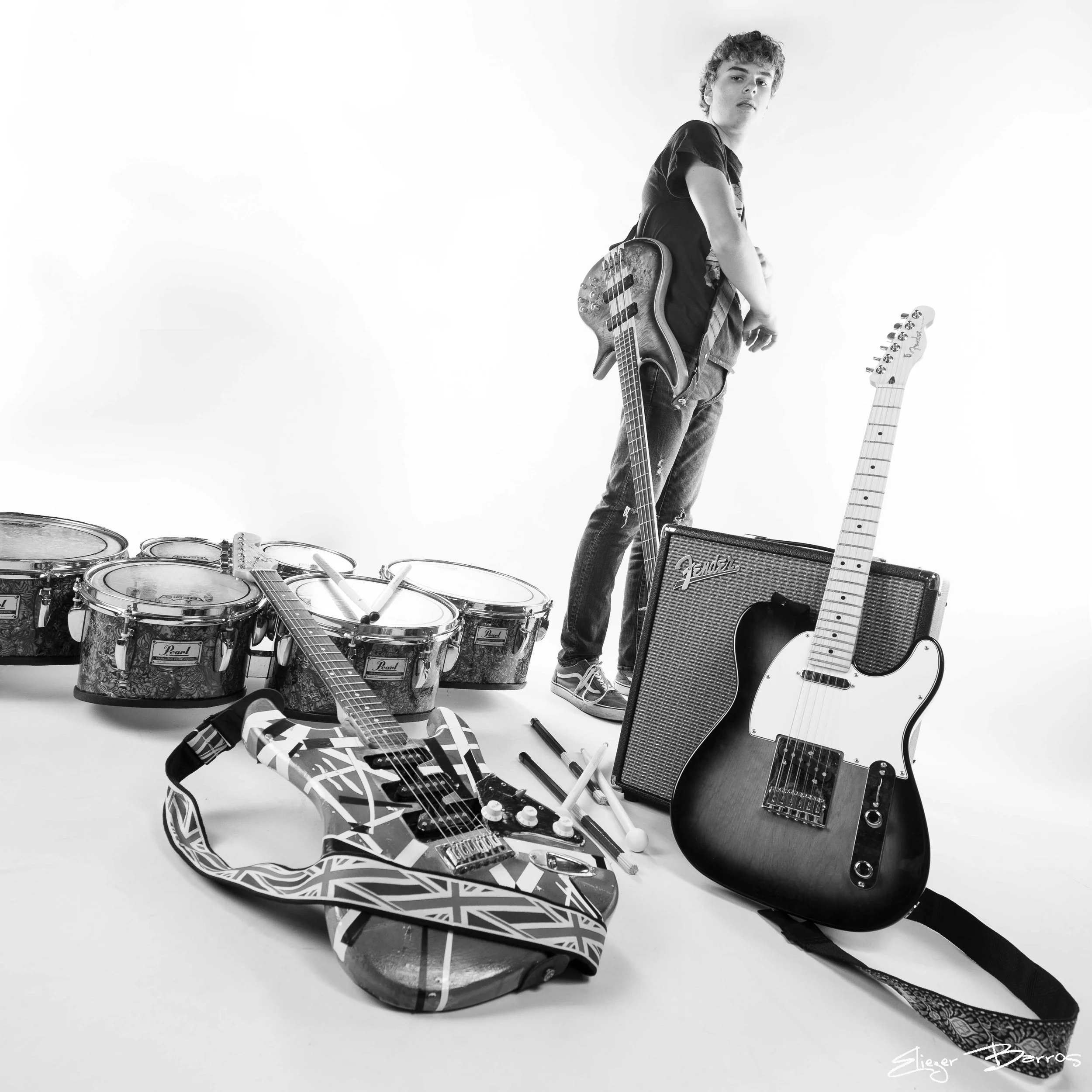 A black and white photo of a young man with curly hair holding an electric bass guitar. There are various musical instruments around him, including a drum set and an acoustic guitar. The background is plain and minimalistic.