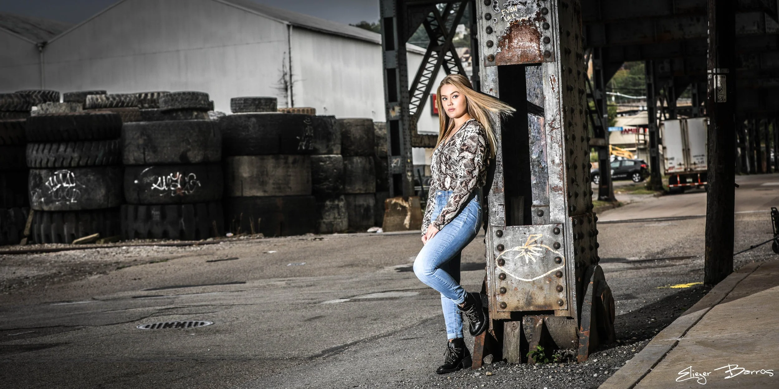 Senior Pictures Pittsburgh in the Strip District. Young woman with blonde hair in a patterned top and jeans leaning against industrial metal structure for bold urban senior portraits.