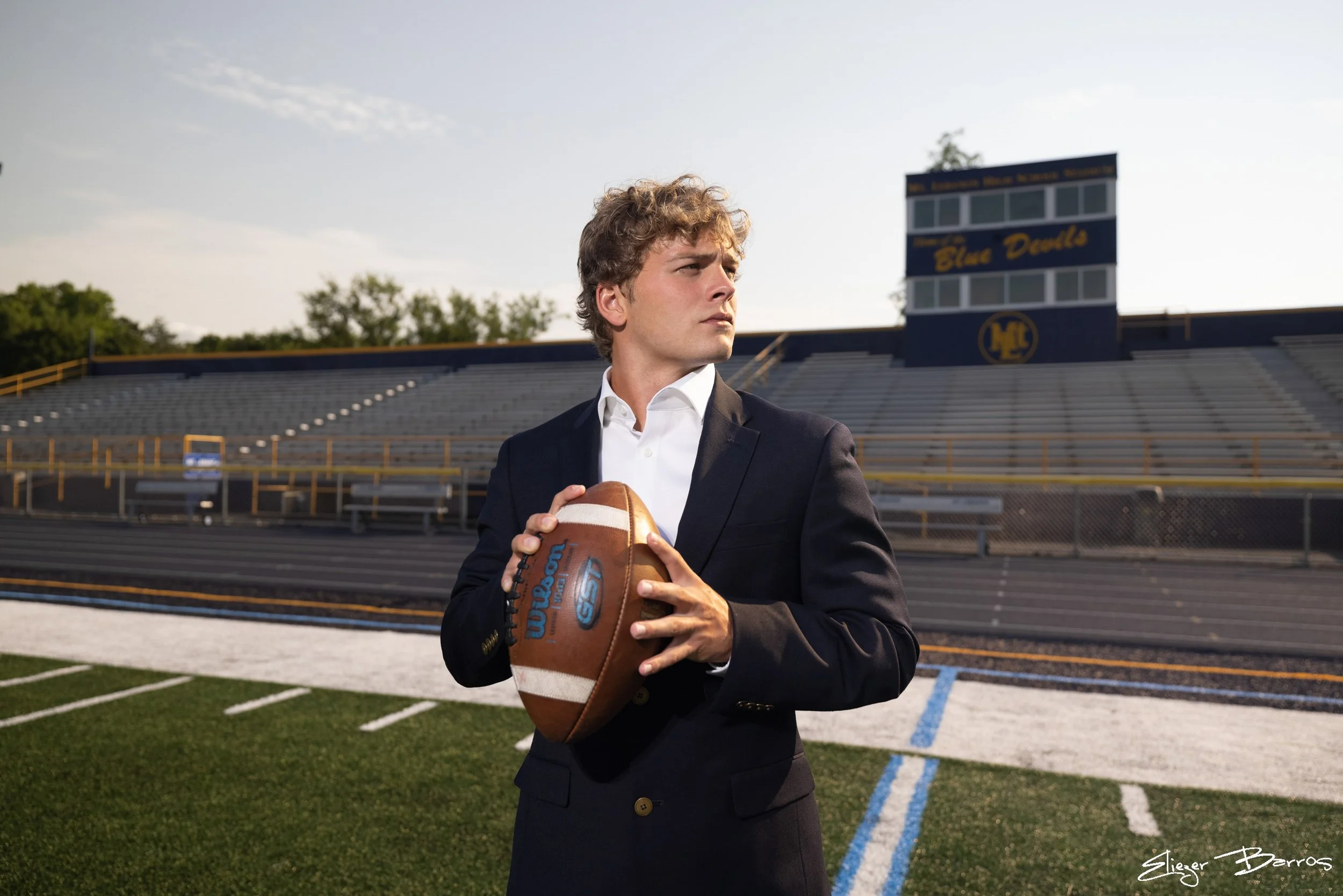 A young man in a black suit and white shirt holding a football on a football field with bleachers and a scoreboard in the background.