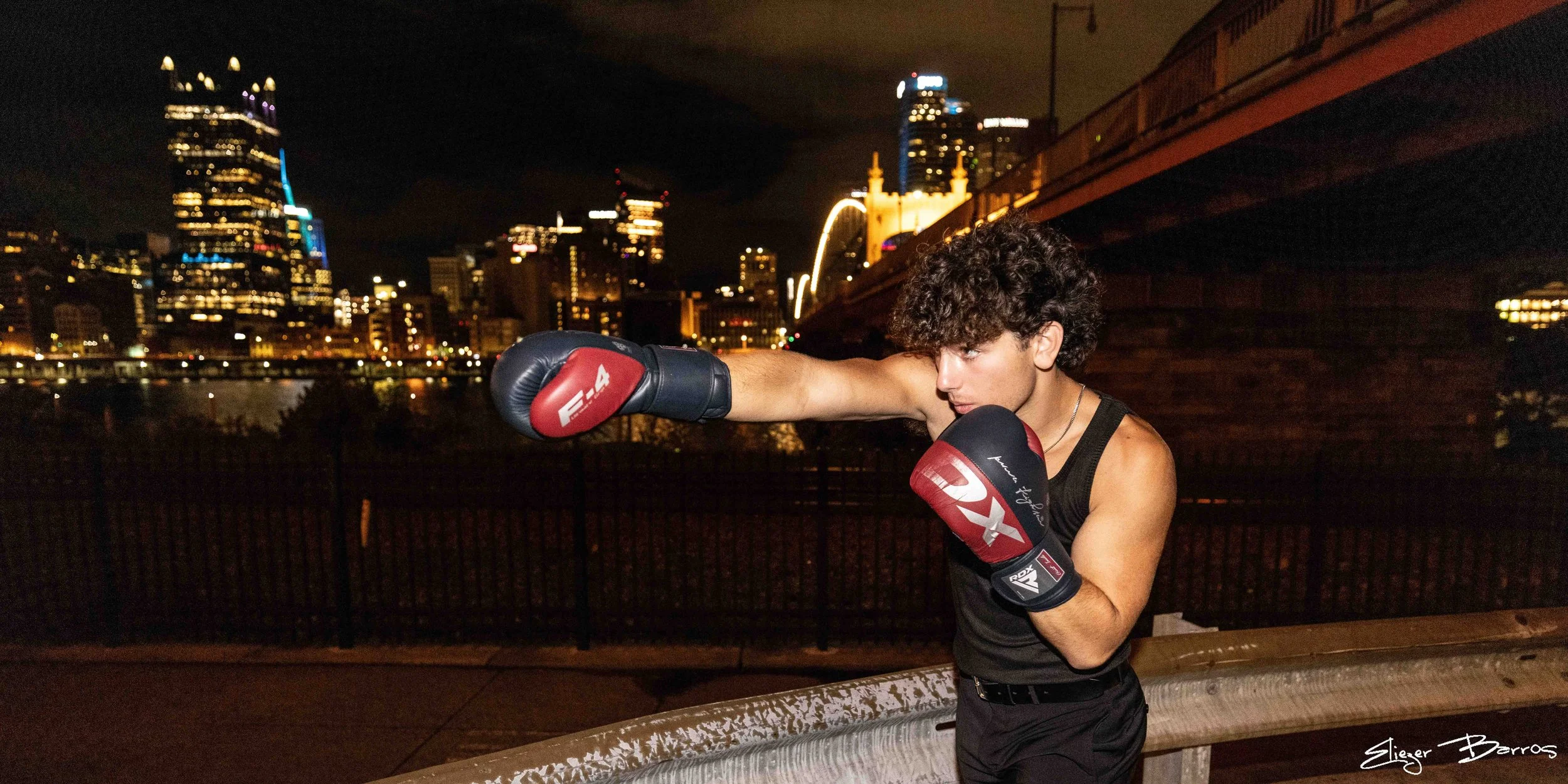 A young woman wearing boxing gloves training outdoors at night with a city skyline illuminated in the background.