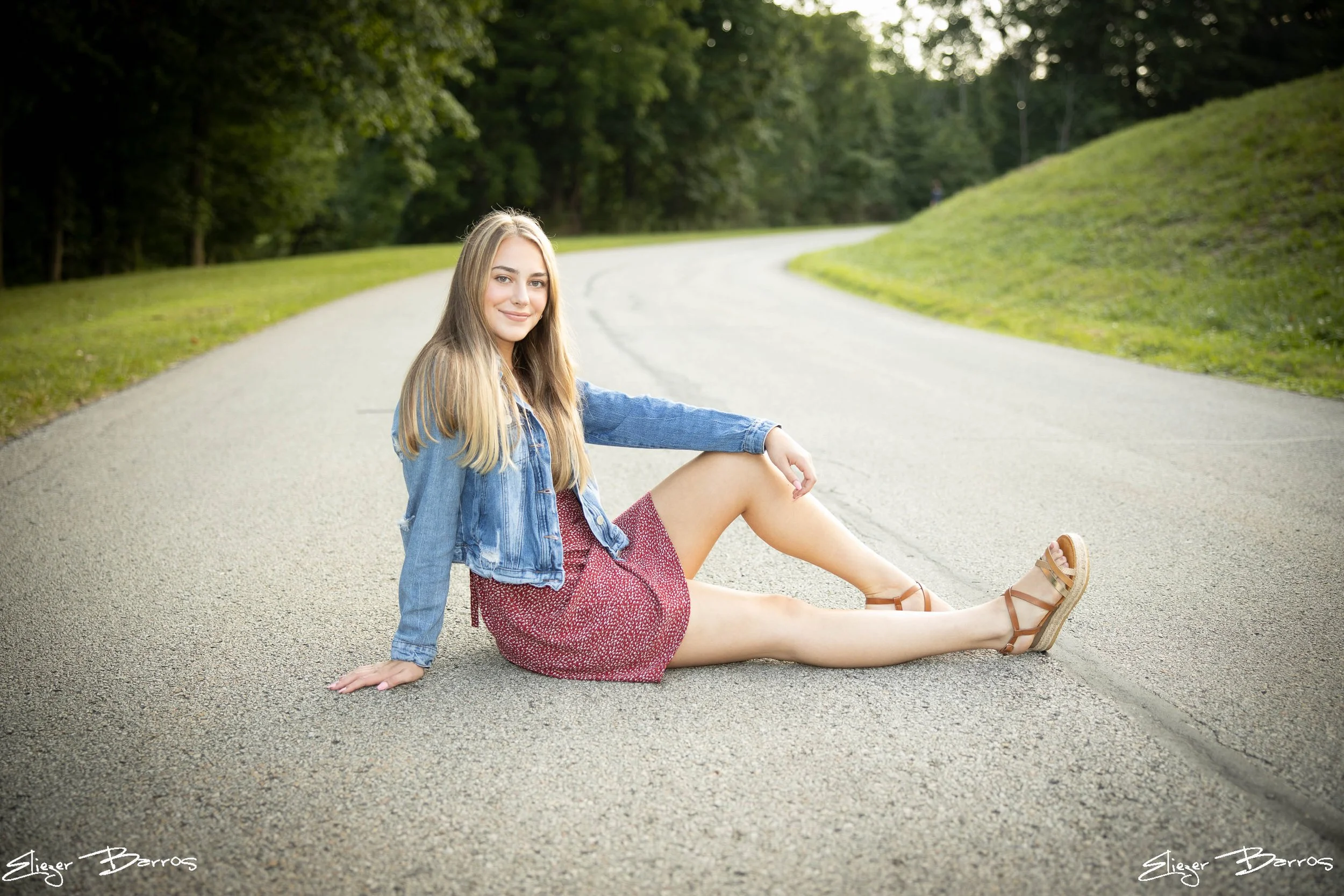 A young woman with long blonde hair sitting on a deserted paved road surrounded by green trees. She is smiling at the camera, wearing a denim jacket, a red patterned dress, and sandals. She is sitting with her legs extended, one hand on the ground an