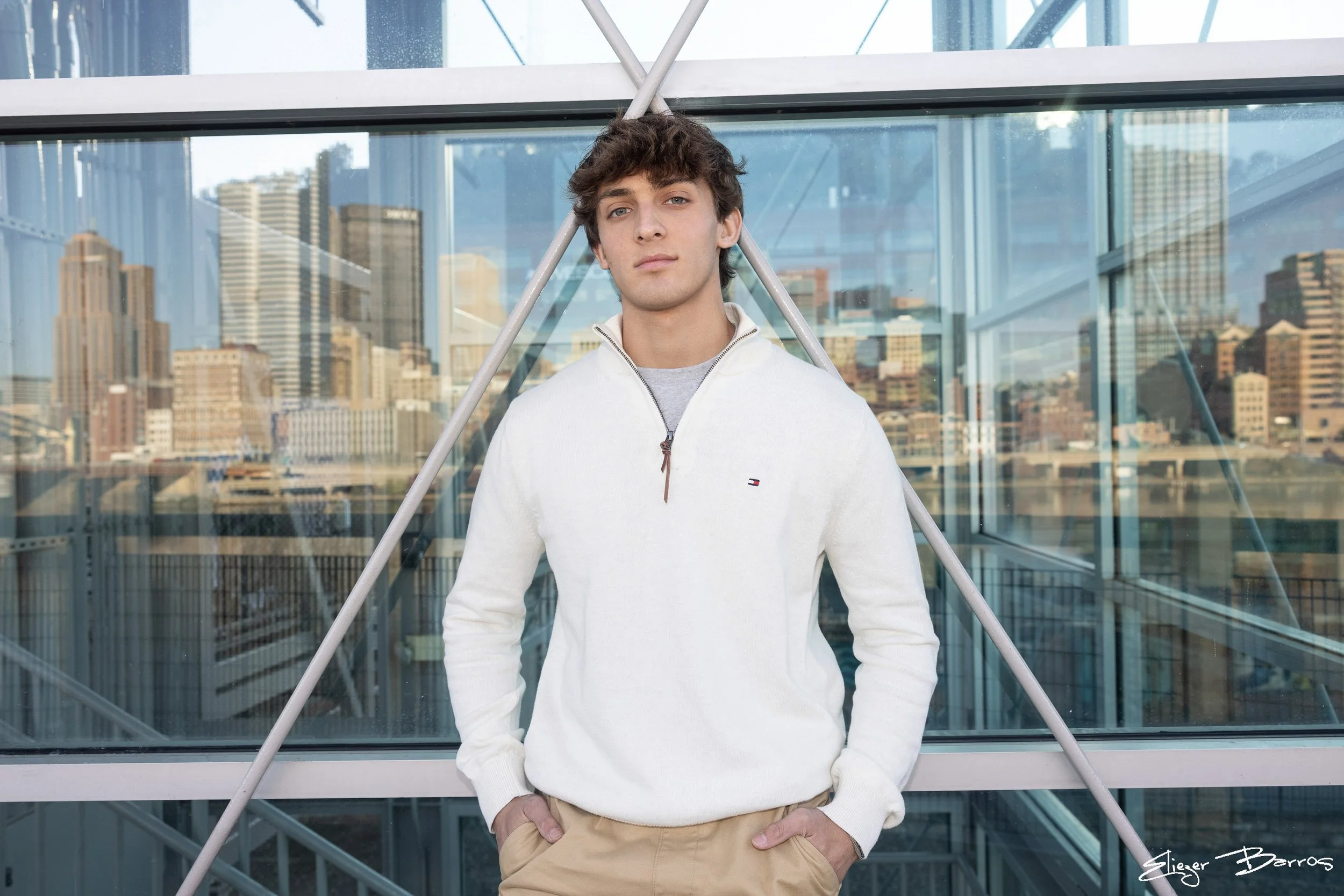 A young man with brown hair and blue eyes standing inside a modern glass building with a city skyline in the background. He is wearing a white zip-up sweater and beige pants, with one hand in his pocket.