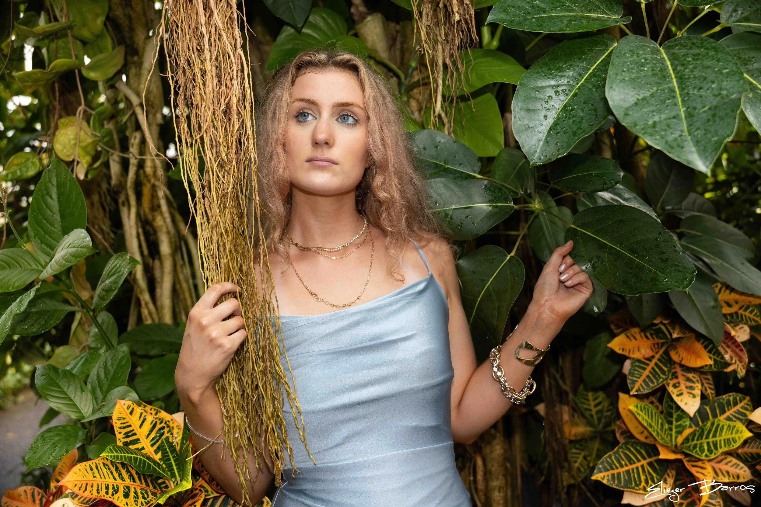 A woman with curly blonde hair and blue eyes standing among green tropical plants. She wears a light blue satin dress, multiple necklaces, and gold jewelry. She is looking to the side with her right hand holding a vine and her left hand touching a le