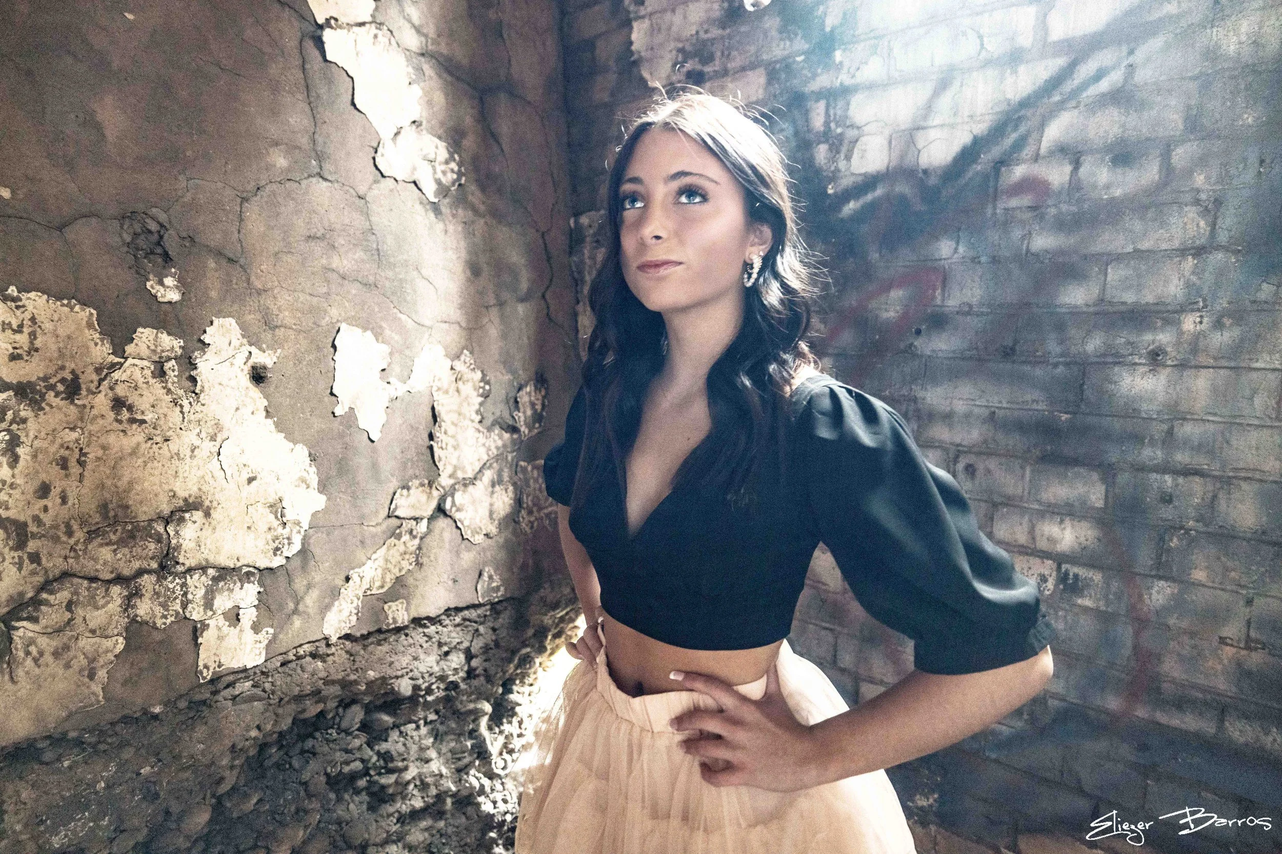 A woman with dark hair and blue eyes, wearing earrings, a black top, and a peach-colored skirt, stands inside an old, weathered brick and concrete structure with peeling paint and exposed bricks. She is looking slightly upward with a soft smile.