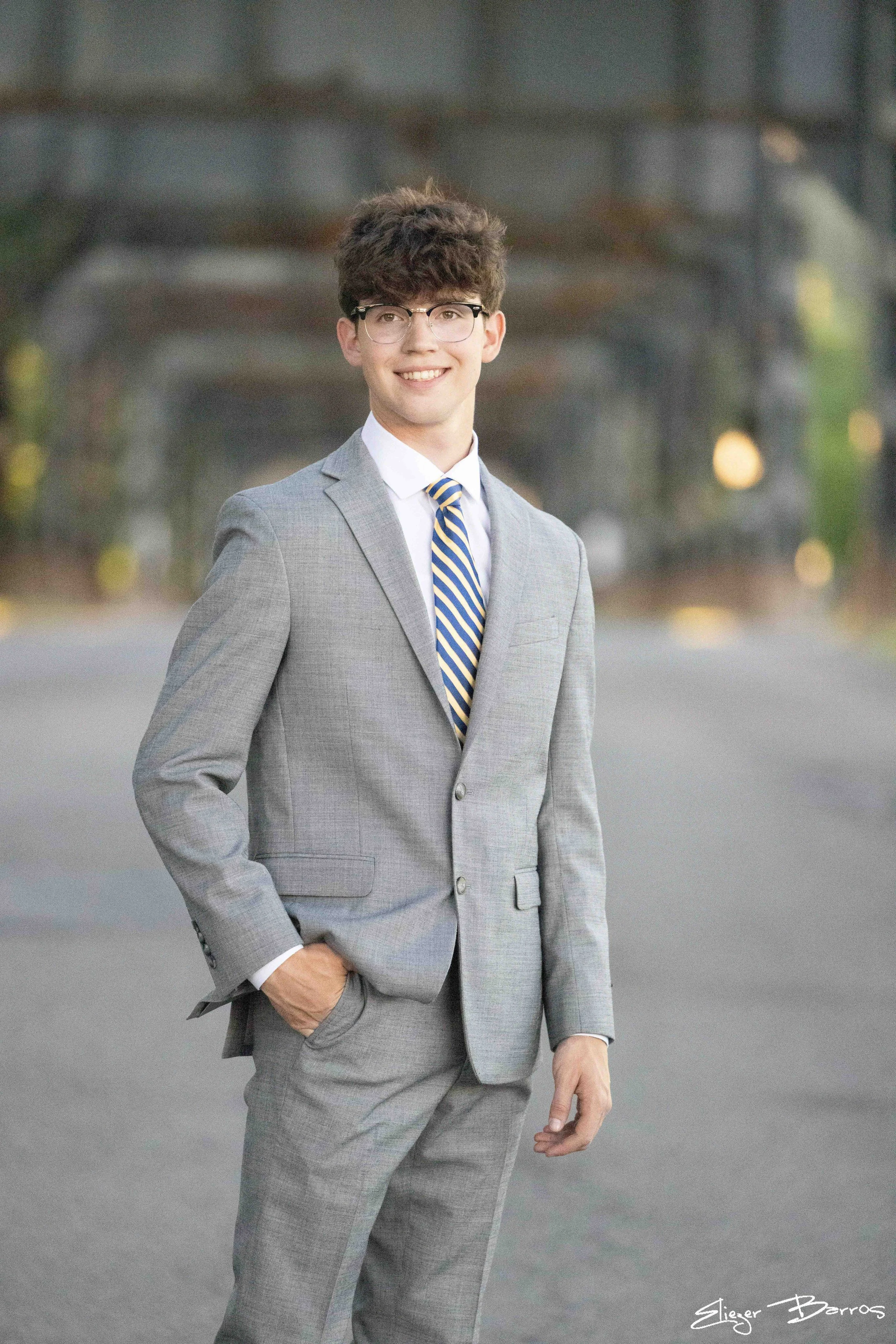 A young man in a gray suit with a white shirt and striped tie, smiling and standing outdoors with blurred trees in the background.