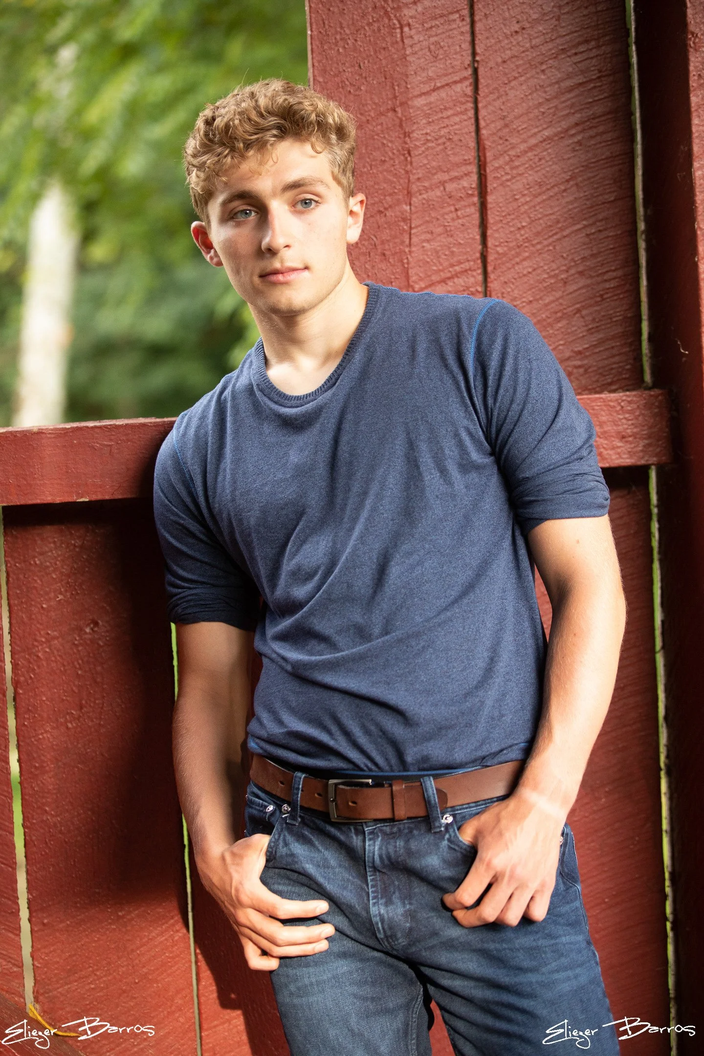 A young man with curly blond hair and blue eyes leaning against a red wooden fence outdoors, wearing a navy blue T-shirt and dark jeans.