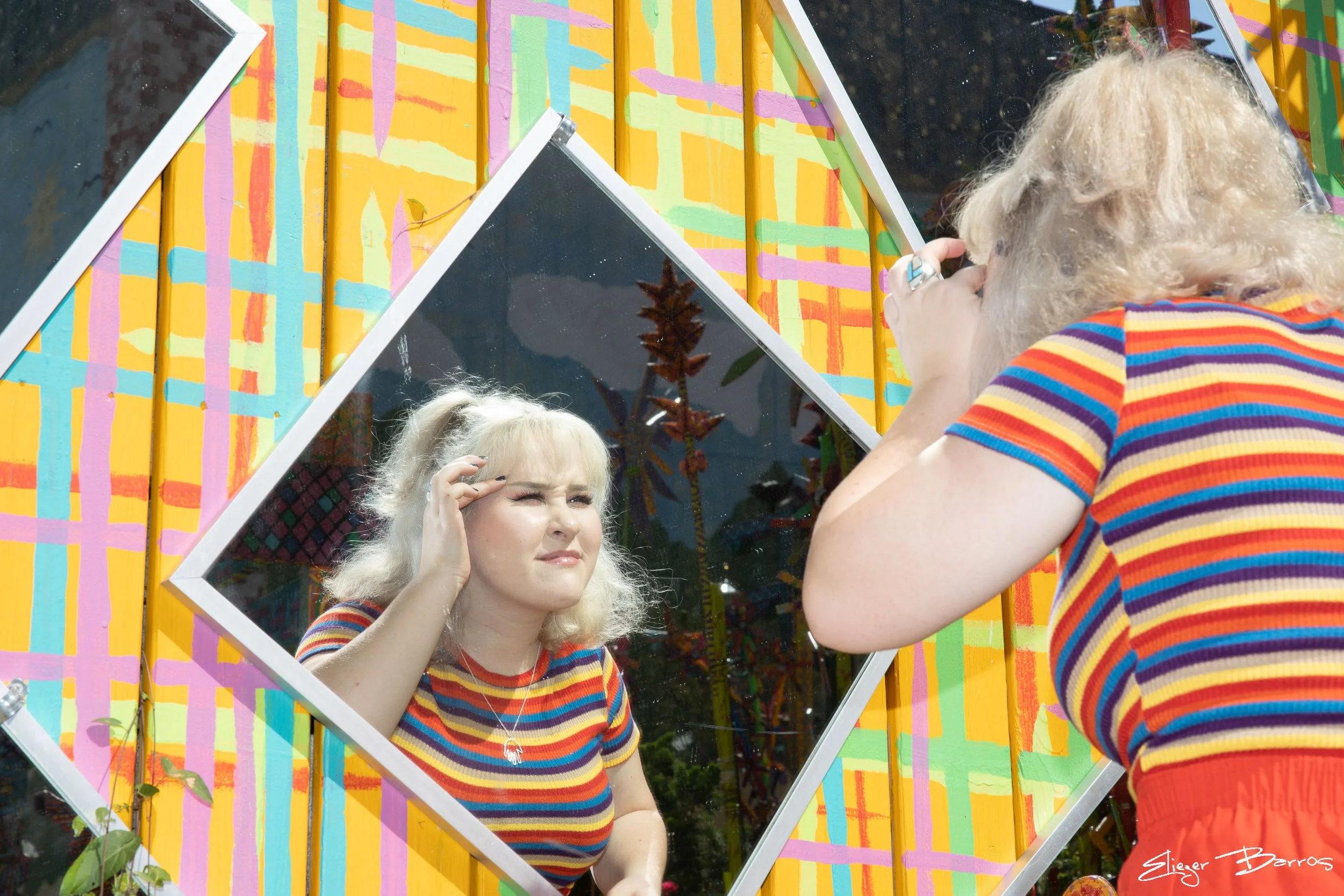 A person with curly blonde hair and a colorful striped shirt is looking into a mirror, adjusting their earrings. The mirror is part of a colorful, geometric outdoor art installation.