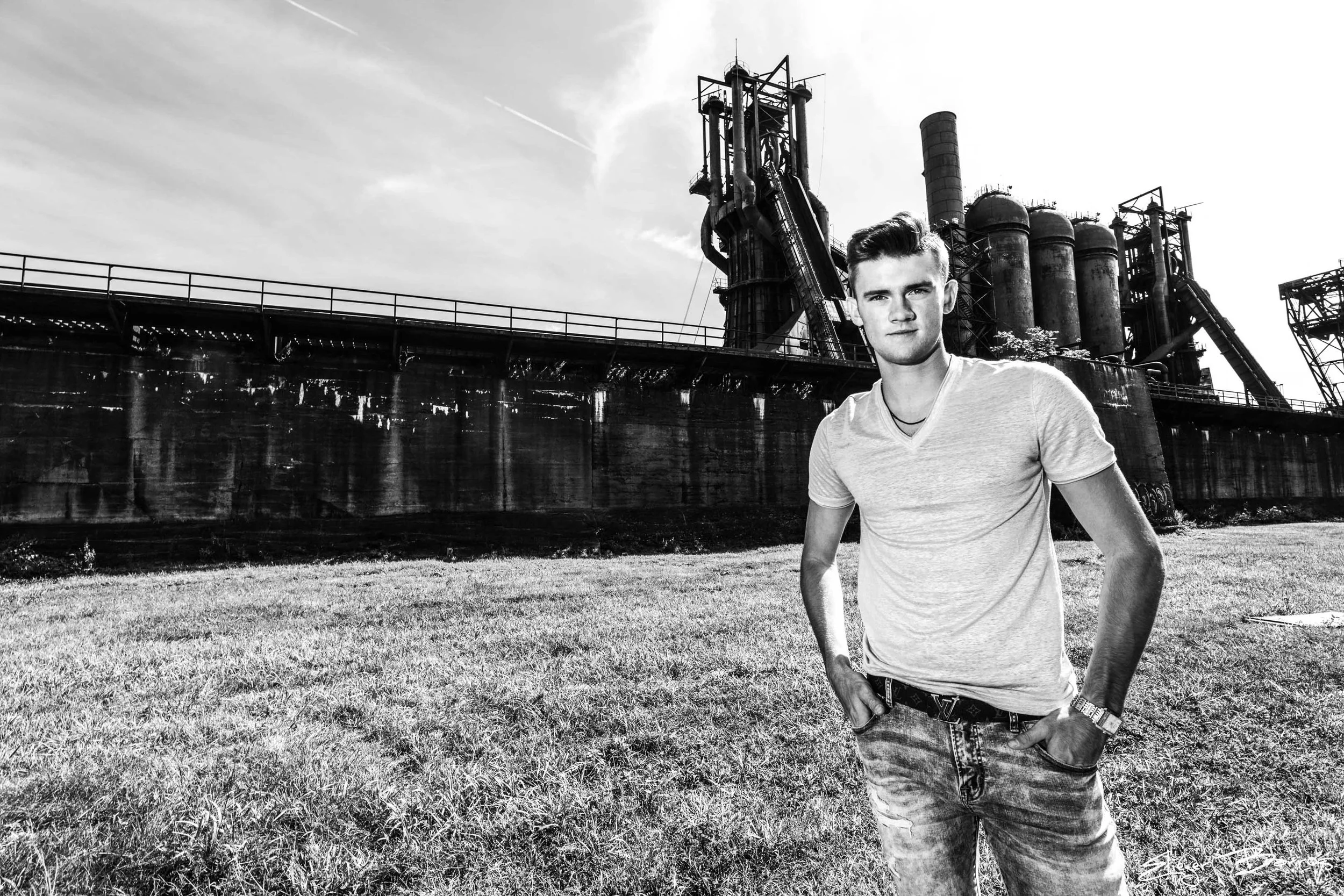 A young man in a light t-shirt and jeans standing on a grassy area with an industrial factory in the background, black and white photo.
