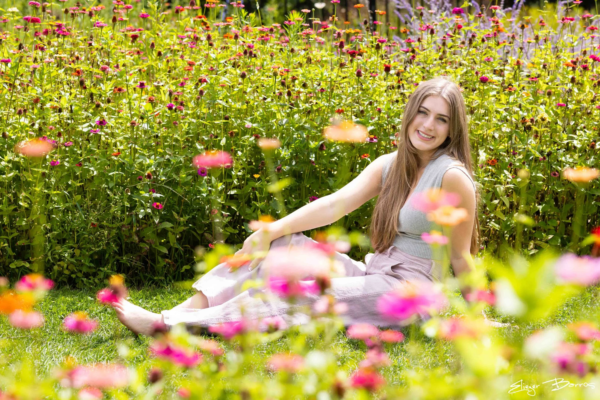 A young woman sitting in a colorful flower garden, smiling and looking at the camera.