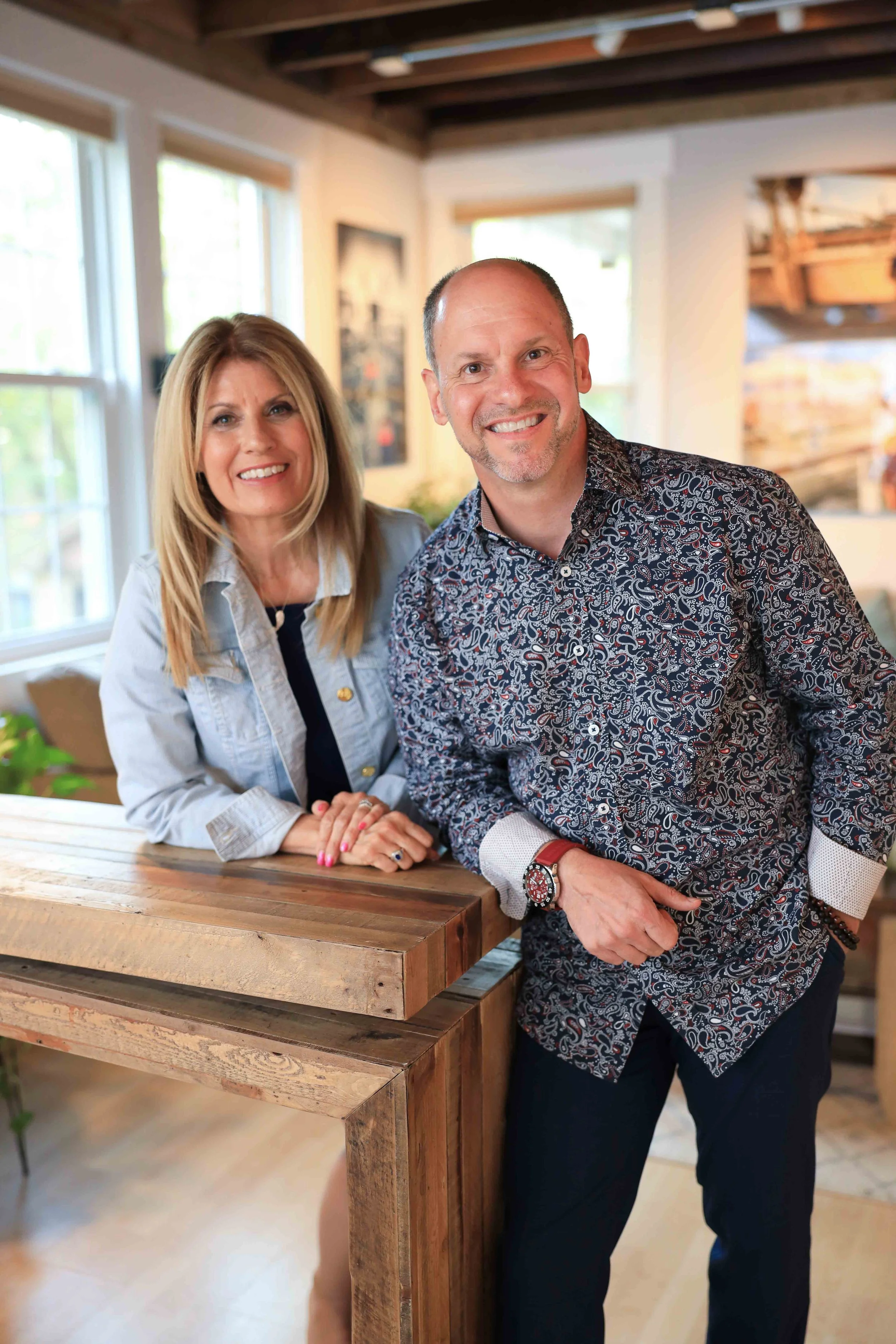 A woman with blonde hair and a man with short dark hair and facial stubble, both smiling, standing indoors near a wooden table. The woman is wearing a light-colored jacket and the man is wearing a patterned dress shirt. The background features window