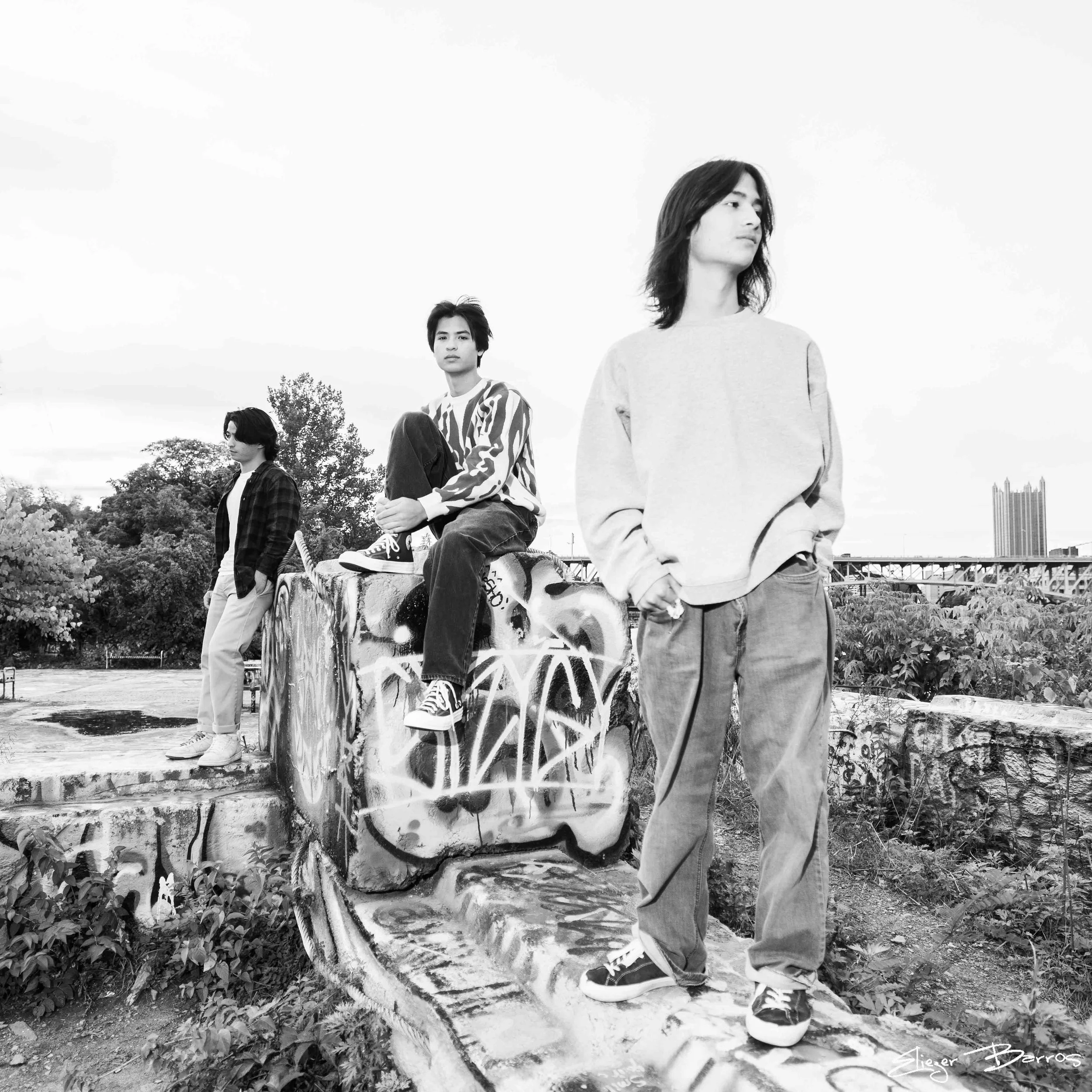 Four young people in casual clothing, one sitting on a graffiti-covered concrete block and the others standing nearby, outdoors with trees and a city skyline in the background, in black and white.