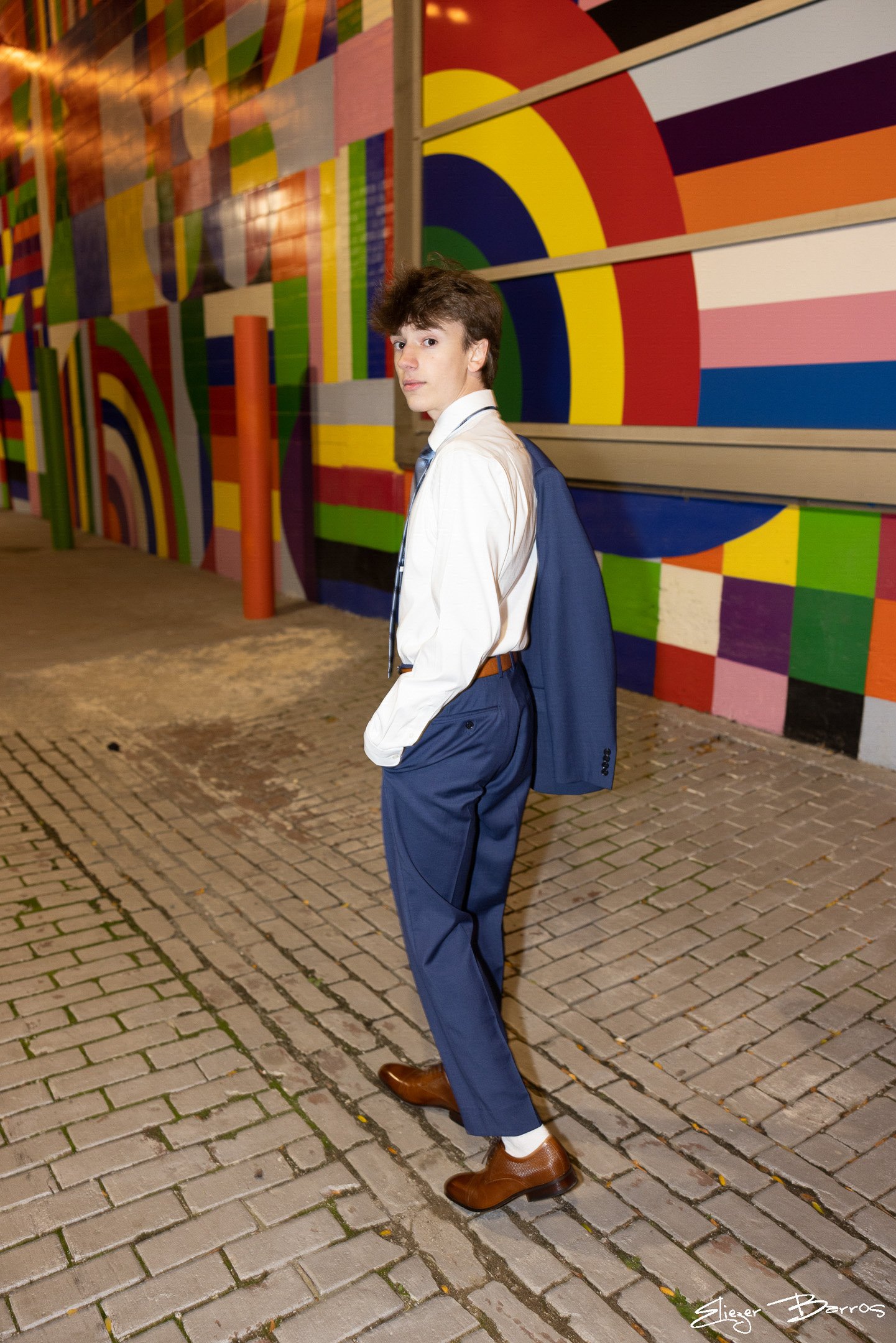 A young man in a white shirt, blue pants, and brown shoes standing on a brick sidewalk with colorful geometric mural art on the wall behind him.