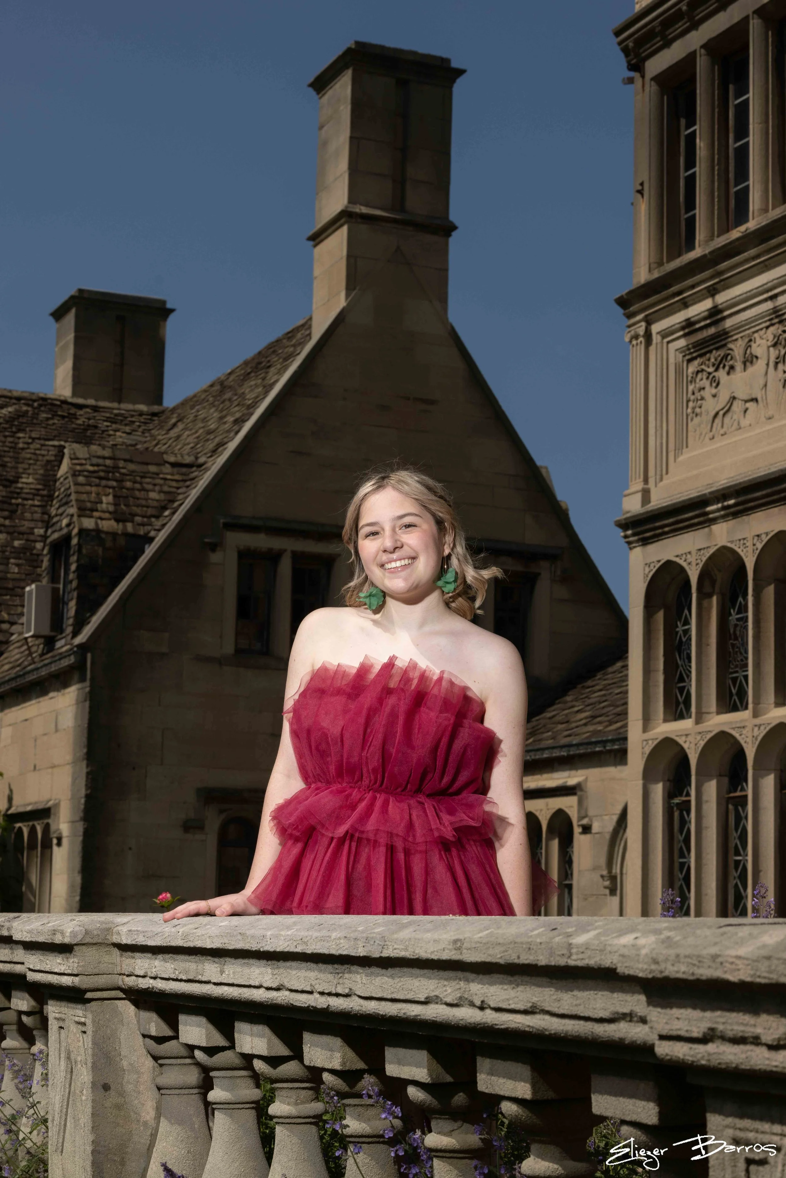 A smiling young woman wearing a strapless red tulle dress with green earrings, standing outdoors against an old stone building with chimneys, arched windows, and decorative stone carvings, under a clear blue sky.