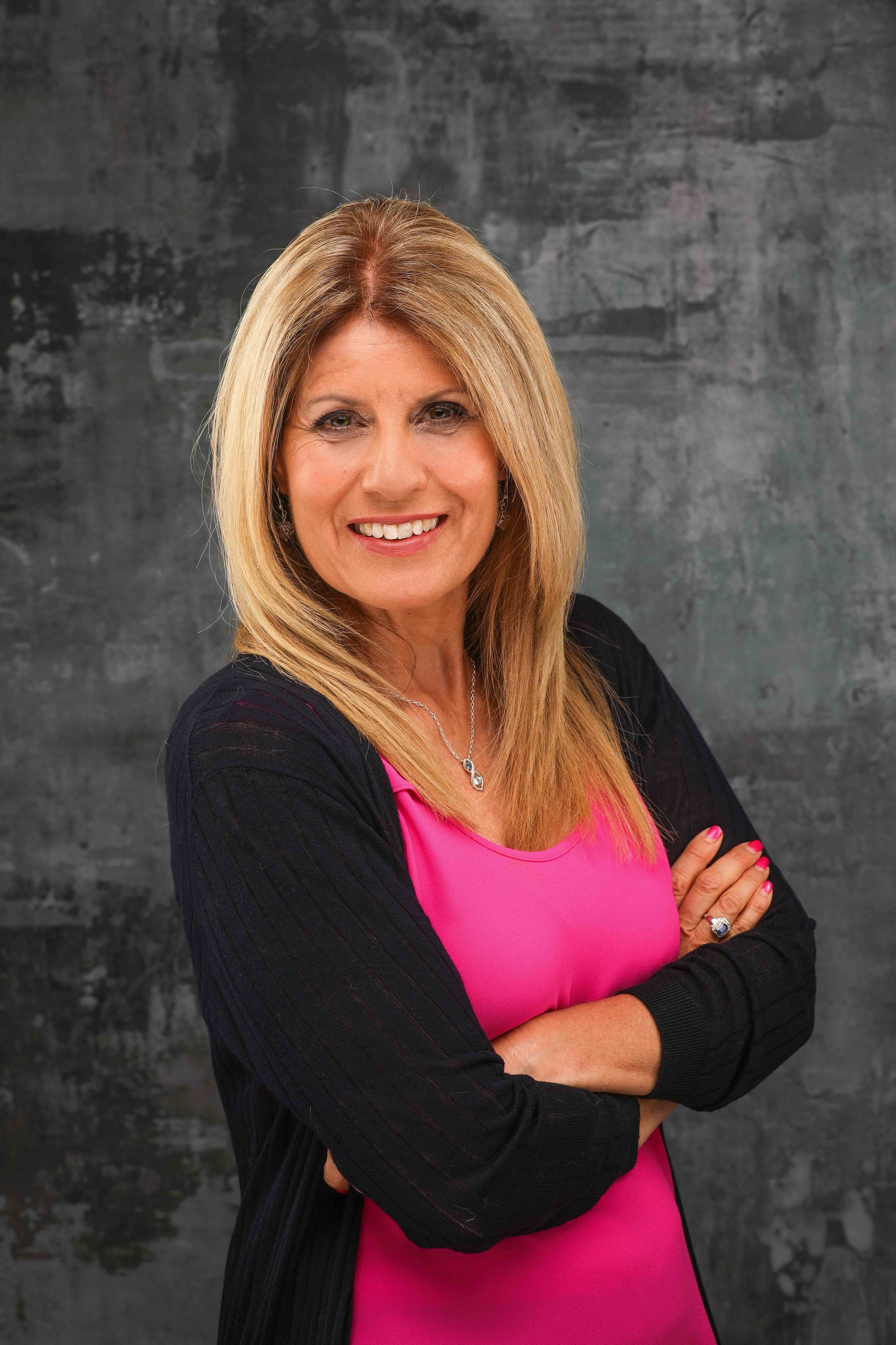 Portrait of a smiling woman with blonde hair, wearing a pink top and black cardigan, standing with arms crossed against a dark textured background.