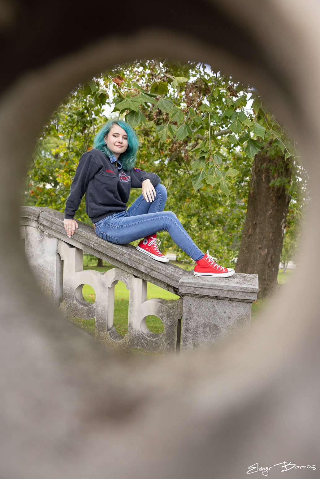 A young woman with teal hair sitting on a stone railing outdoors, viewed through a circular opening with leaves and trees in the background.