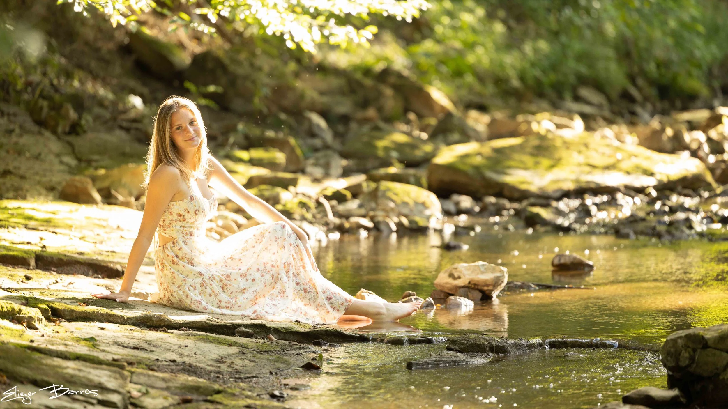 Senior Pictures Pittsburgh at Mingo Creek Park. Young woman in a flowing floral dress relaxing on rocks by the creek near the historic Ebenezer Covered Bridge for peaceful natural outdoor senior portraits.