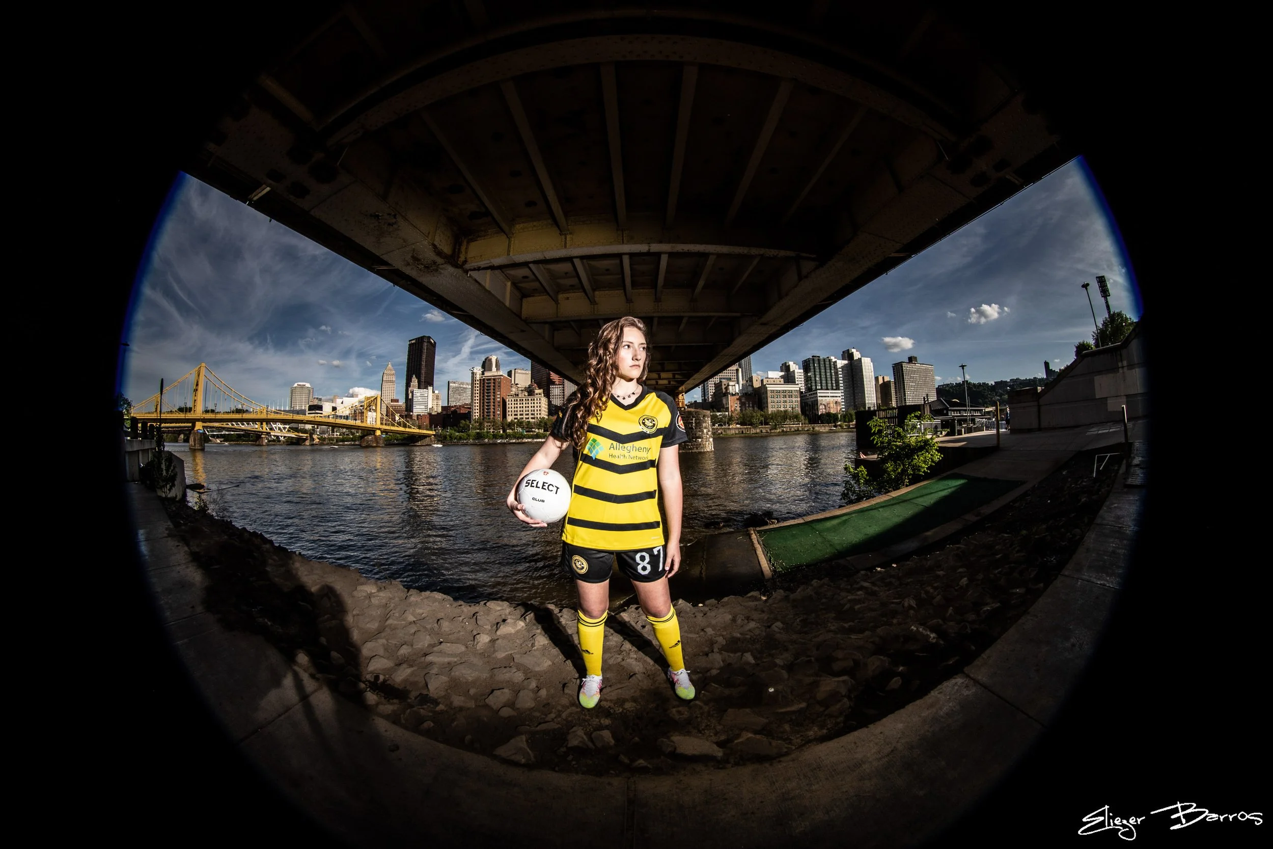 A young woman in a yellow and black soccer uniform standing under a bridge, holding a soccer ball, with a city skyline and a river in the background.