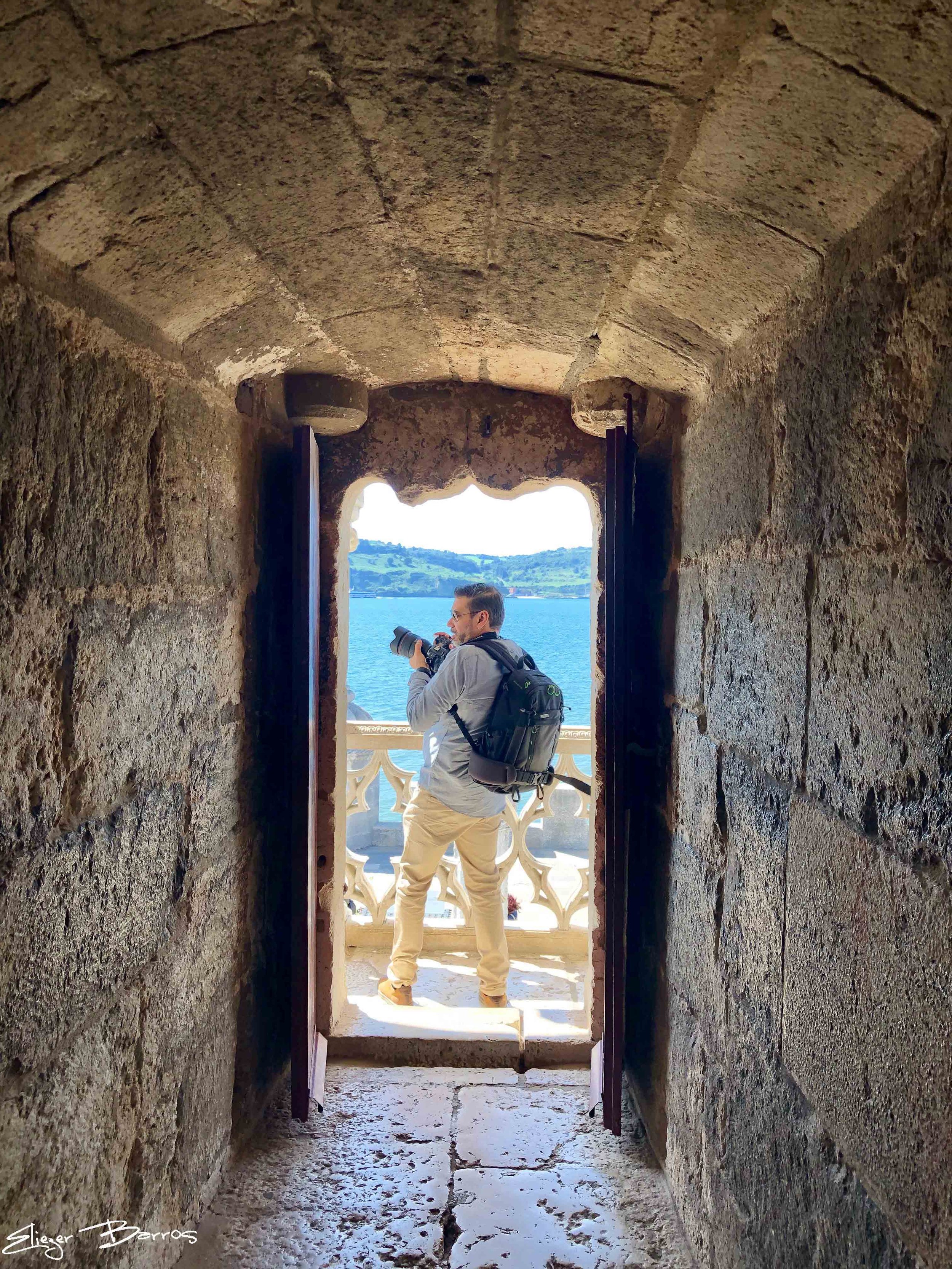 A man with glasses, wearing a gray shirt and beige pants, stands on a balcony with a white patterned railing, looking at water and hills in the background, viewed through a stone archway.