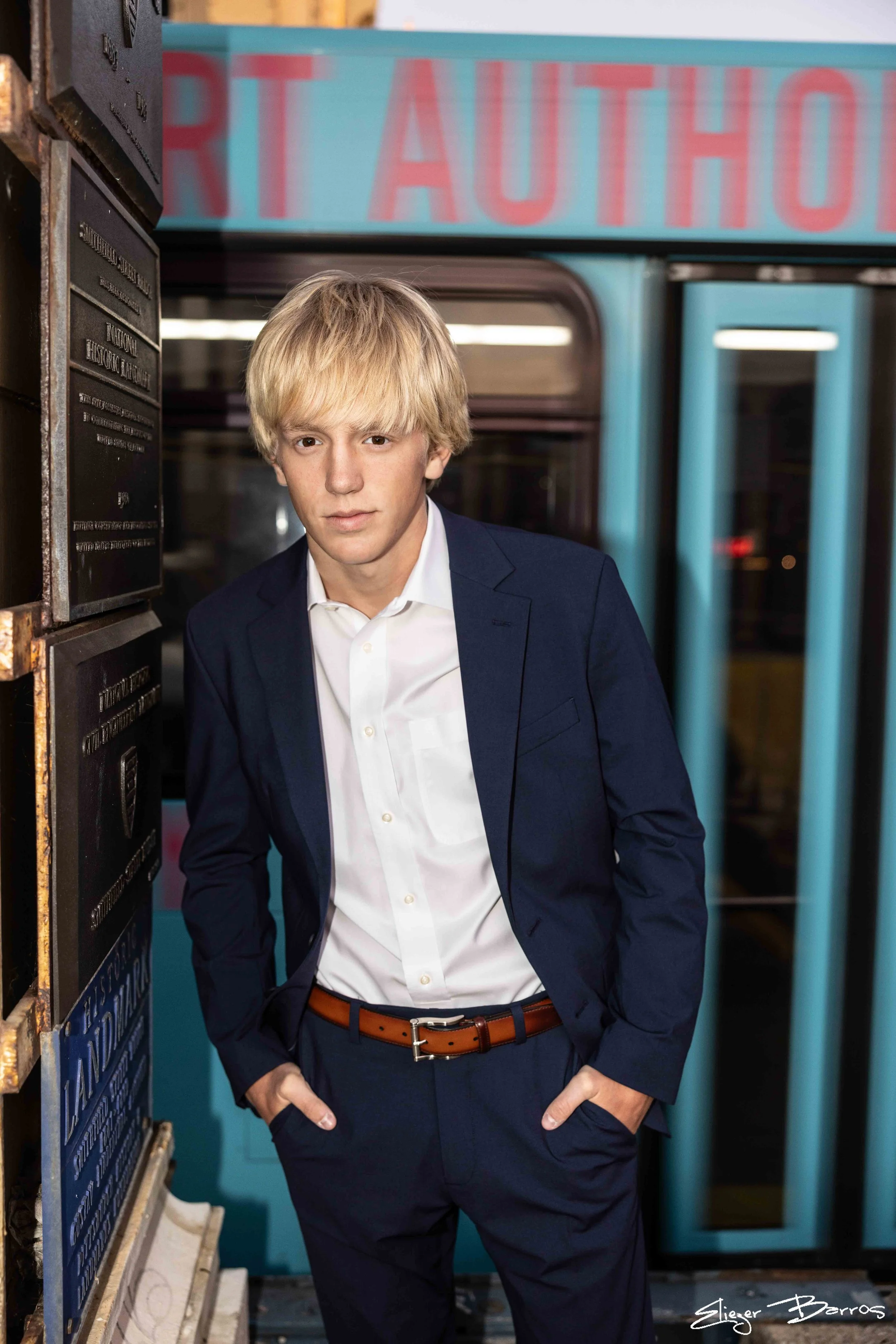 A young man with blonde hair wearing a dark blue suit and white shirt standing in front of a blue streetcar with red lettering, hands in his pockets, in an urban environment.