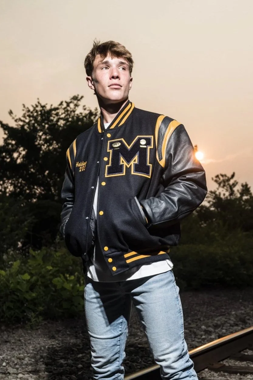 Senior Pictures Pittsburgh at Hot Metal Bridge. Young man in a black and gold varsity jacket posing with hands in pockets on the historic bridge at sunset for stylish modern senior portraits.